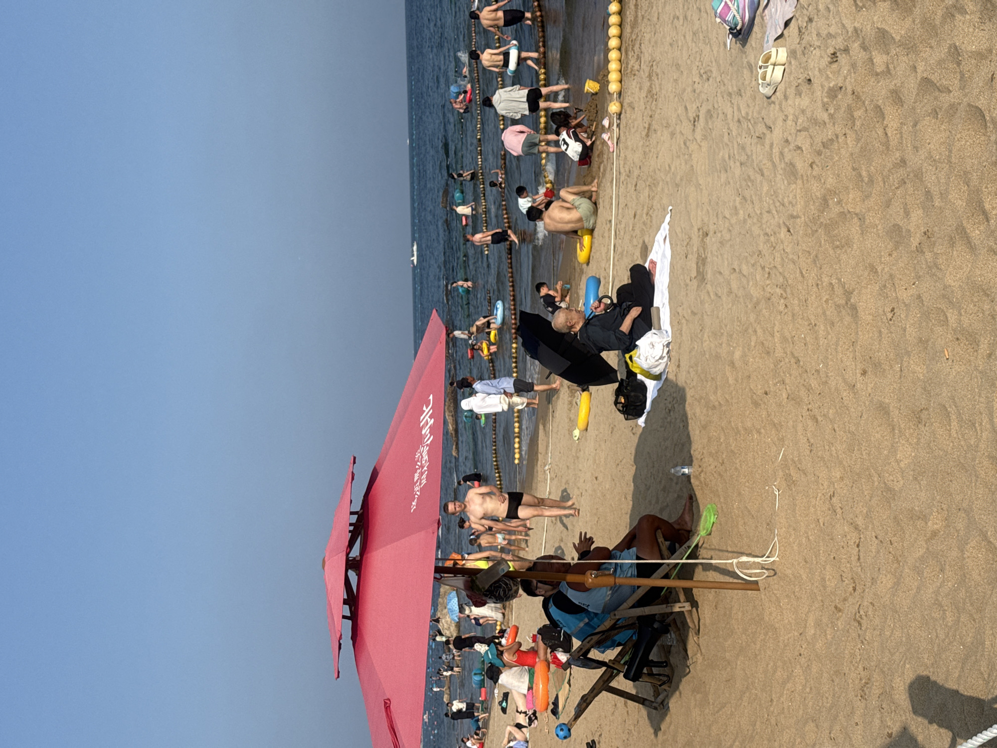 The image depicts a crowded beach scene in Qinhuangdao, China. The beach is sandy with a calm sea in the background. Many people are visible, some wading in the shallow water, others sitting or playing on the sand. A large red umbrella is prominently positioned on the left side of the frame, providing shade. Several beach chairs are also visible. People are dressed in swimwear and casual beach attire. The water is cordoned off with buoys and ropes, indicating a designated swimming area. The sky is clear and blue. The overall impression is of a lively, recreational day at the beach.