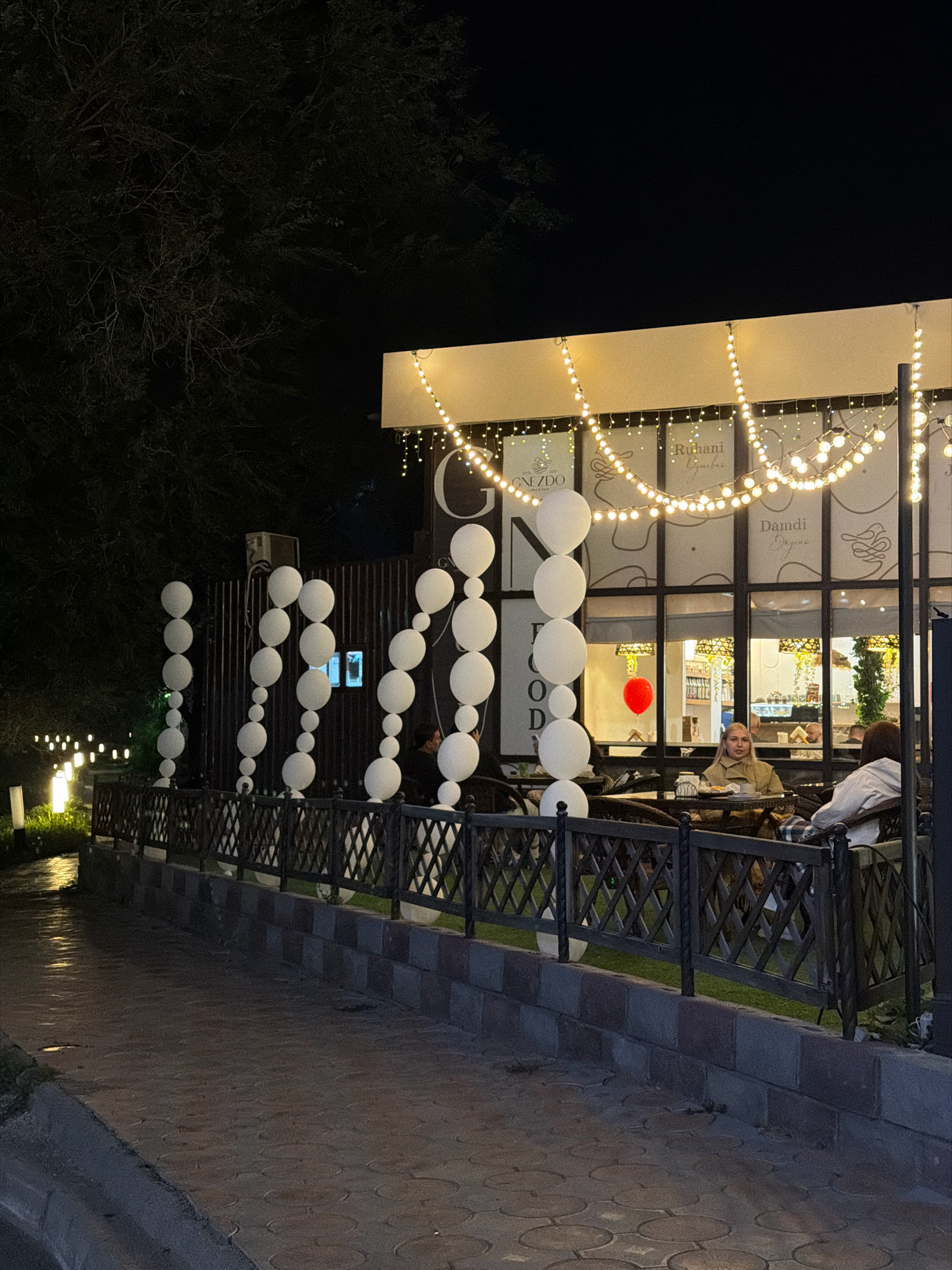 This image depicts an outdoor seating area of a cafe or restaurant at night, possibly located in Pavlodar, Kazakhstan. The scene is illuminated by string lights and pathway lights. In the foreground, a paved walkway runs alongside a dark metal fence. Beyond the fence, there is a strip of green grass and tables with chairs where patrons are seated.
Several large white balloons are arranged in vertical columns along the fence line, serving as decoration. One of the patrons is a young woman with blonde hair, seated at a table. Behind her, another person wearing a white hoodie is visible. The building behind the seating area has large windows, through which the interior of the establishment can be seen, displaying shelves and lights. A red balloon is visible hanging inside the building. The overall ambiance is that of a casual, lit-up gathering spot at night.