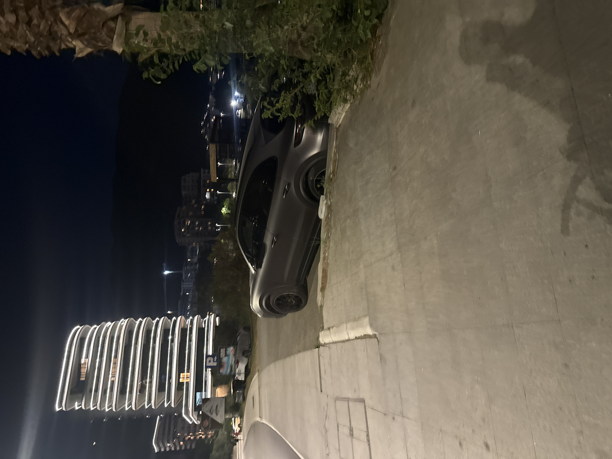 In Boreti, Montenegro, a dark grey Porsche Cayenne SUV is parked on a curb next to a sidewalk at night. The vehicle's rear tires are partially on the sidewalk and appears to be stuck. In the background, a modern, illuminated skyscraper with a wavy facade is visible, along with other buildings and lights of the city. A palm tree with green foliage is present on the right side of the image. The sky is dark, suggesting it is nighttime.