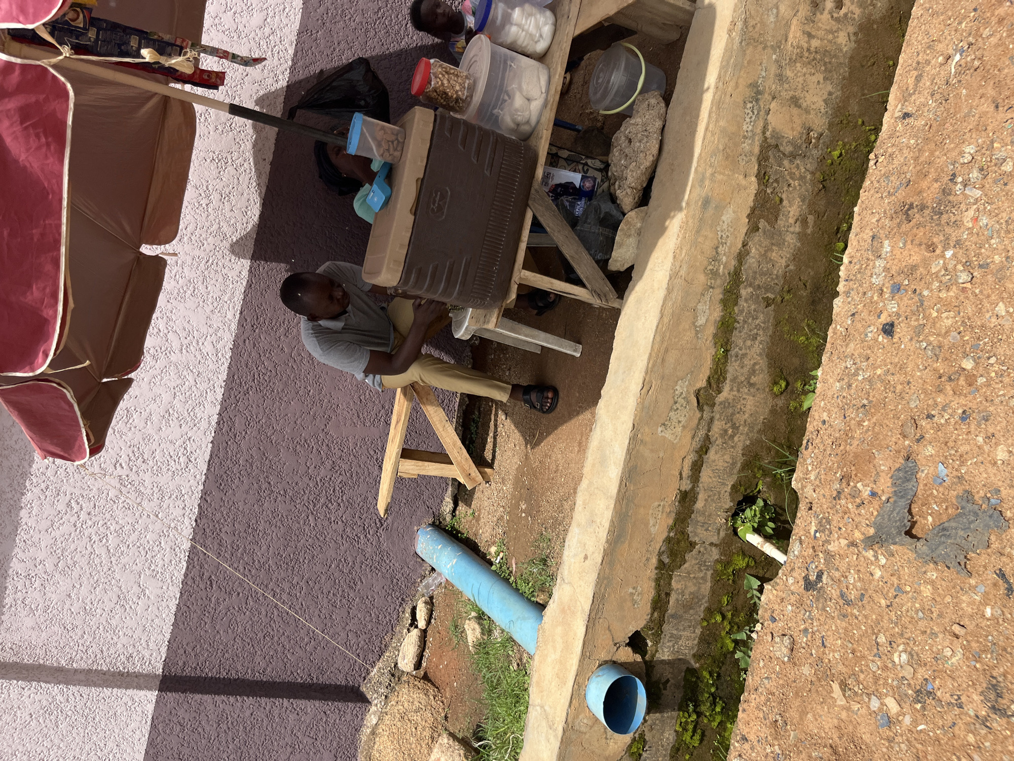 A man sits on a wooden bench under a large umbrella in an outdoor market. He is wearing a gray polo shirt and tan pants, and is looking down at something in his lap. Next to him, a woman is lying down. On a table in front of them are several containers of food, including a large brown cooler. A blue pipe runs along the ground behind them. The scene appears to be located in Bukuru, Nigeria.