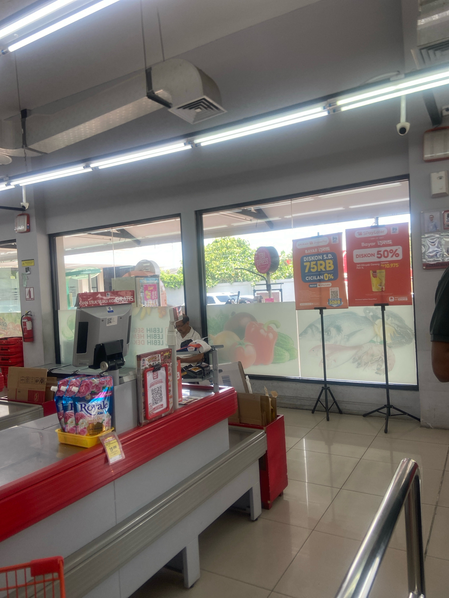This is an interior shot of a supermarket checkout area in Ngaglik, Indonesia. A cashier, an adult male wearing a white polo shirt, is visible behind the counter, facing away from the camera. The checkout counter is made of a red and grey material. To the left of the cashier, there are several products, including a blue bag of Royale fabric softener. A red fire extinguisher is mounted on a pillar to the left of the frame, near a stack of red baskets. Above the counter, a computer monitor is positioned for the cashier. In the background, large windows look out onto a sunny outdoor area with greenery. Two promotional posters are displayed in front of the windows. The floor is tiled in a light beige color, and fluorescent lighting illuminates the ceiling.