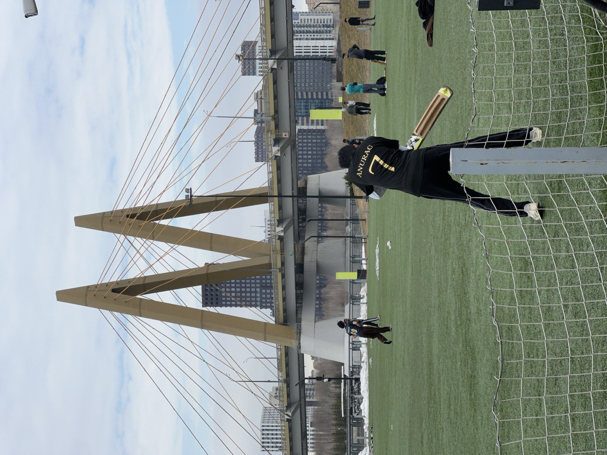 The image shows a cricket game taking place near the Millennium Bridge in Kazan, Russia. A person wearing a black jersey with the name "ANURAG" and the number "7" is holding a cricket bat. Several other people are visible in the background. There's a green field, a bridge in the background, and buildings. A net is on the right of the frame. The sky is cloudy.