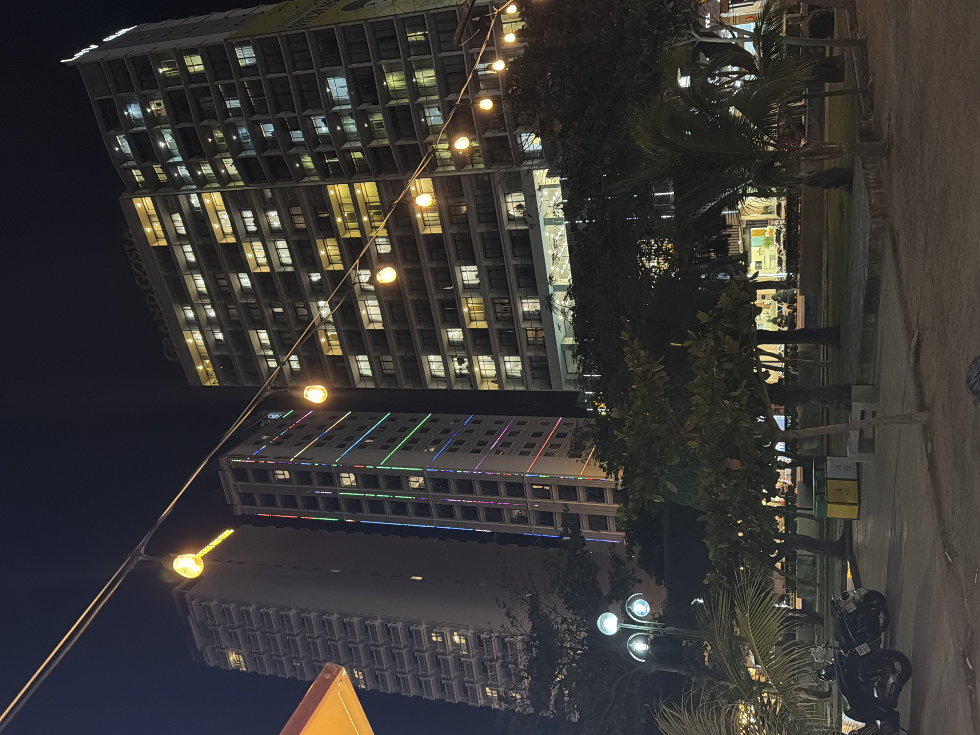 This nighttime outdoor scene features three high-rise buildings in Nha Trang Ward, Vietnam. The tallest building on the left has a name on the side that reads "GRAND COSTA". Its windows are illuminated with a warm glow. Below this is another high-rise with colorful lines of light running vertically along the building. The third high-rise is less illuminated and further away. A string of lights hangs across the scene, blurring some of the details in the background. Trees and other vegetation fill the lower right of the frame. In the foreground, there is a motorbike parked and other unidentified objects. The lighting is dim and artificial.