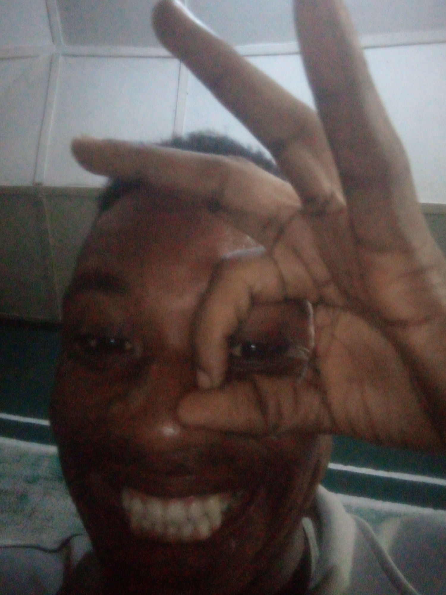 A person with dark skin is smiling and holding their hand in front of one eye, forming a circle with their thumb and index finger. Two other fingers are extended upwards. The background appears to be an interior setting, possibly with tiled walls. The location is Unknown, Unknown.