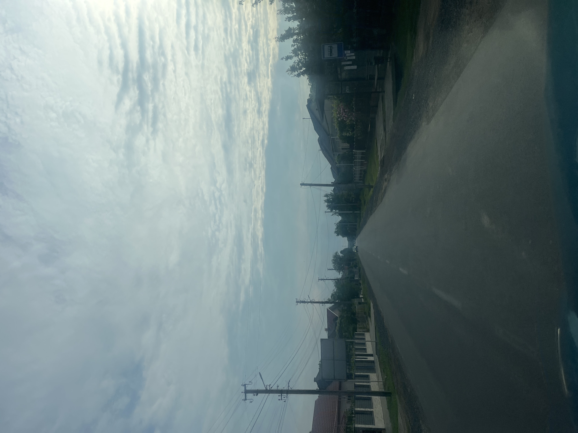 The image presents a roadside view in Storozhnytsia, Ukraine, likely captured from a moving vehicle. A paved road stretches into the distance, appearing dark grey and slightly reflective in the foreground, with a lighter grey or white dividing line visible further down the road.

Along the left side of the road, a series of residential houses are visible, featuring pitched roofs and fences, often partially obscured by trees and other vegetation. Numerous utility poles, primarily wooden, line the road, carrying multiple strands of overhead power lines that crisscross the sky. Lush green trees and bushes are abundant, contributing to a verdant landscape alongside the dwellings.

The sky occupies a significant portion of the frame, filled with scattered, streaky clouds that suggest a partly cloudy day. The overall lighting is bright but diffused, indicating late morning or early afternoon with the sun likely behind the clouds or positioned higher in the sky.

A prominent blue and white bus stop sign, featuring a white bus icon, is visible on the left side of the road. Below it, several smaller, indistinct signs are attached, with any text illegible due to distance and resolution. In the far distance, a small, light-colored vehicle can be faintly seen on the road. No people are visible, and the scene depicts a quiet, everyday street in a rural or semi-rural setting.