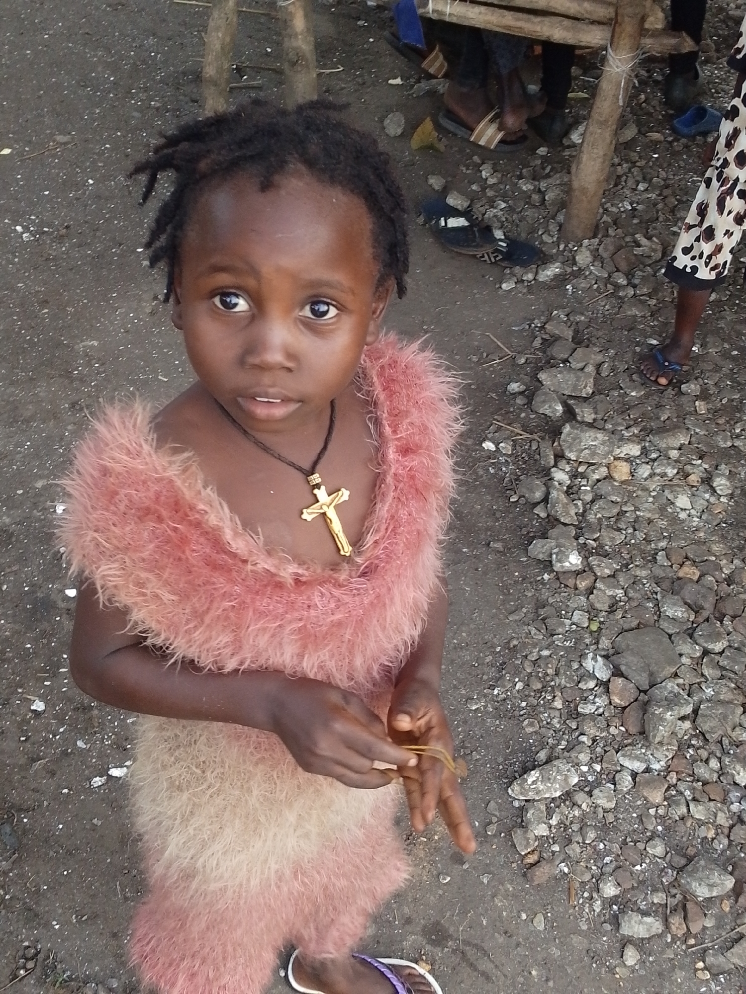 The image shows a young Black child in Otulu, Nigeria, standing in a dirt and gravel area. The child is wearing a pink, fluffy garment that covers their shoulders and torso, and a gold cross necklace. The child's expression is directed towards the camera. In the background, there are glimpses of other people and their feet, some wearing sandals. There are also some wooden structures and scattered debris. The overall environment appears to be an outdoor, possibly informal, setting.