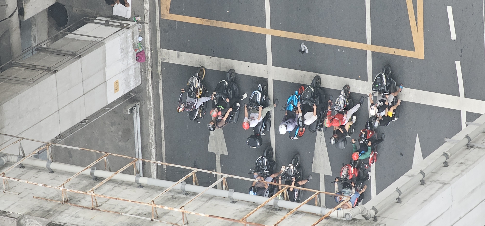 This is an aerial, high-angle photograph taken in the city of Makati, Philippines, depicting a line of motorcycles and their riders on an asphalt road, possibly a pedestrian overpass or an elevated walkway based on the surrounding concrete structure and scaffolding. The motorcycles are parked in a single file, facing towards the top of the image. Several riders are seated on their vehicles, some with passengers. One motorcycle has a blue body, while others are primarily black. The road is marked with white and yellow lines, including a large white arrow indicating direction. In the background, on a raised concrete platform, there are some personal belongings, including a pink bag and several smaller items. The overall scene is one of a momentary pause or waiting period for the motorcyclists.