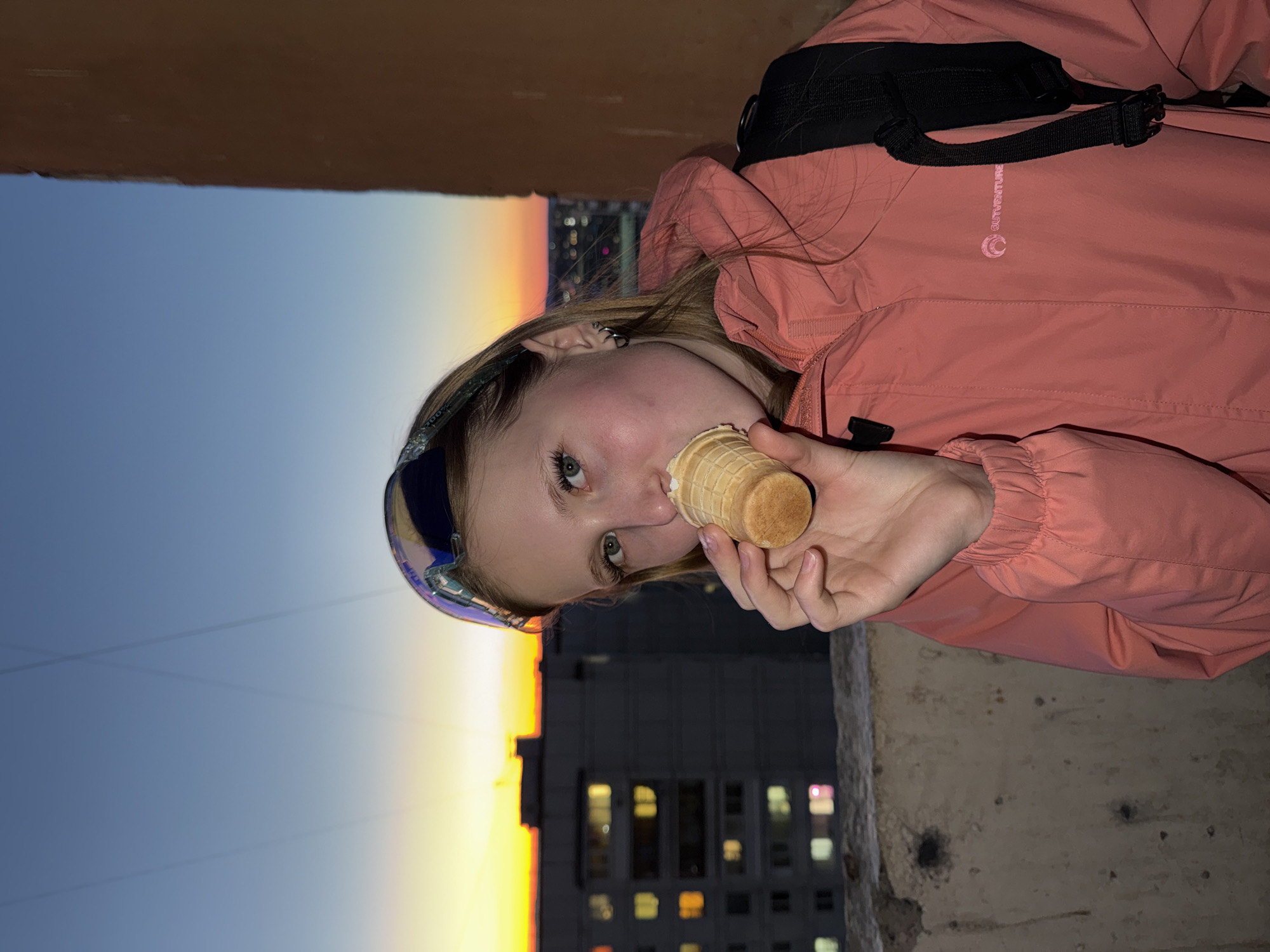 A young woman is eating an ice cream cone. She is wearing mirrored sunglasses on top of her head and a reddish-orange jacket. She has a black backpack over her shoulder. The backdrop includes a building with lit windows and a gradient sky that fades from blue to orange. The photo was taken in Chaikovsky, Russia.