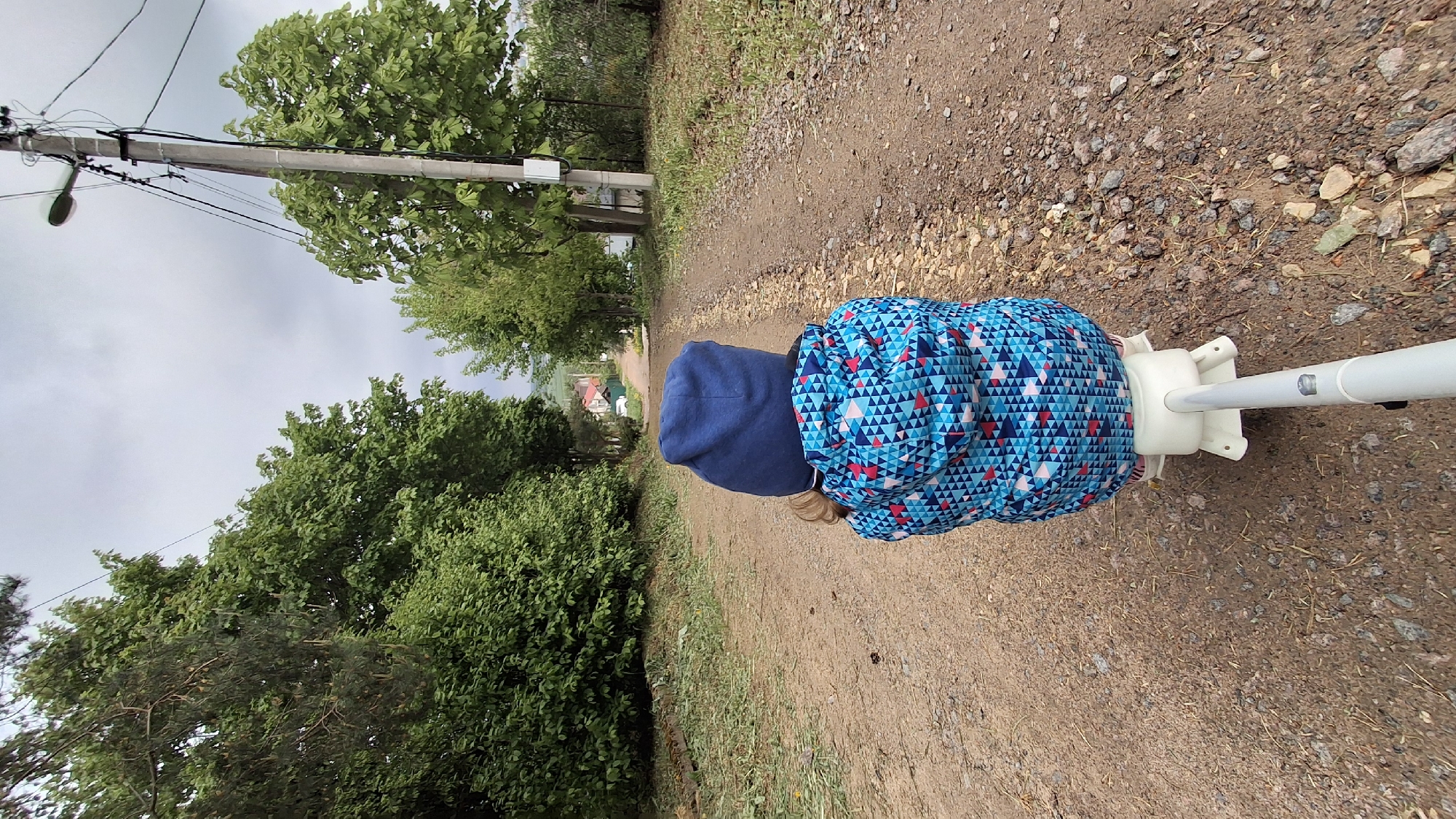A person wearing a blue hooded jacket and a blue jacket with a triangle pattern is walking on a dirt road. The location appears to be Kempelevo, Russia, based on visual information. A white scooter frame is visible near the person. The environment includes trees, foliage, a utility pole, and a visible sky. The scene suggests an outdoor setting on a cloudy day.