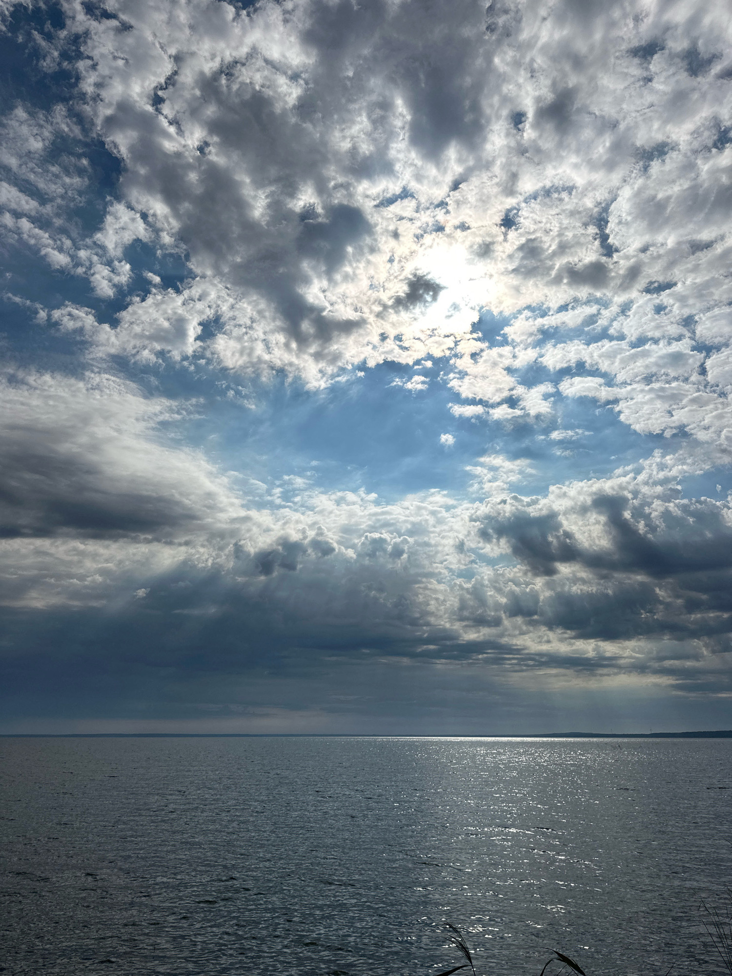 The image depicts a wide, calm body of water under a dramatic sky filled with a mixture of dark and bright clouds. Sunlight filters through gaps in the clouds, creating bright rays that illuminate parts of the sky and reflect off the water's surface, forming a shimmering path. The horizon is clearly visible, with a faint landmass on the distant edge. The foreground shows the dark, rippled surface of the water, with some blurred blades of grass or reeds at the very bottom right corner. The scene is devoid of any people or structures, suggesting a natural, open landscape. The location is identified as Нарацкі сельскі Савет, Belarus.