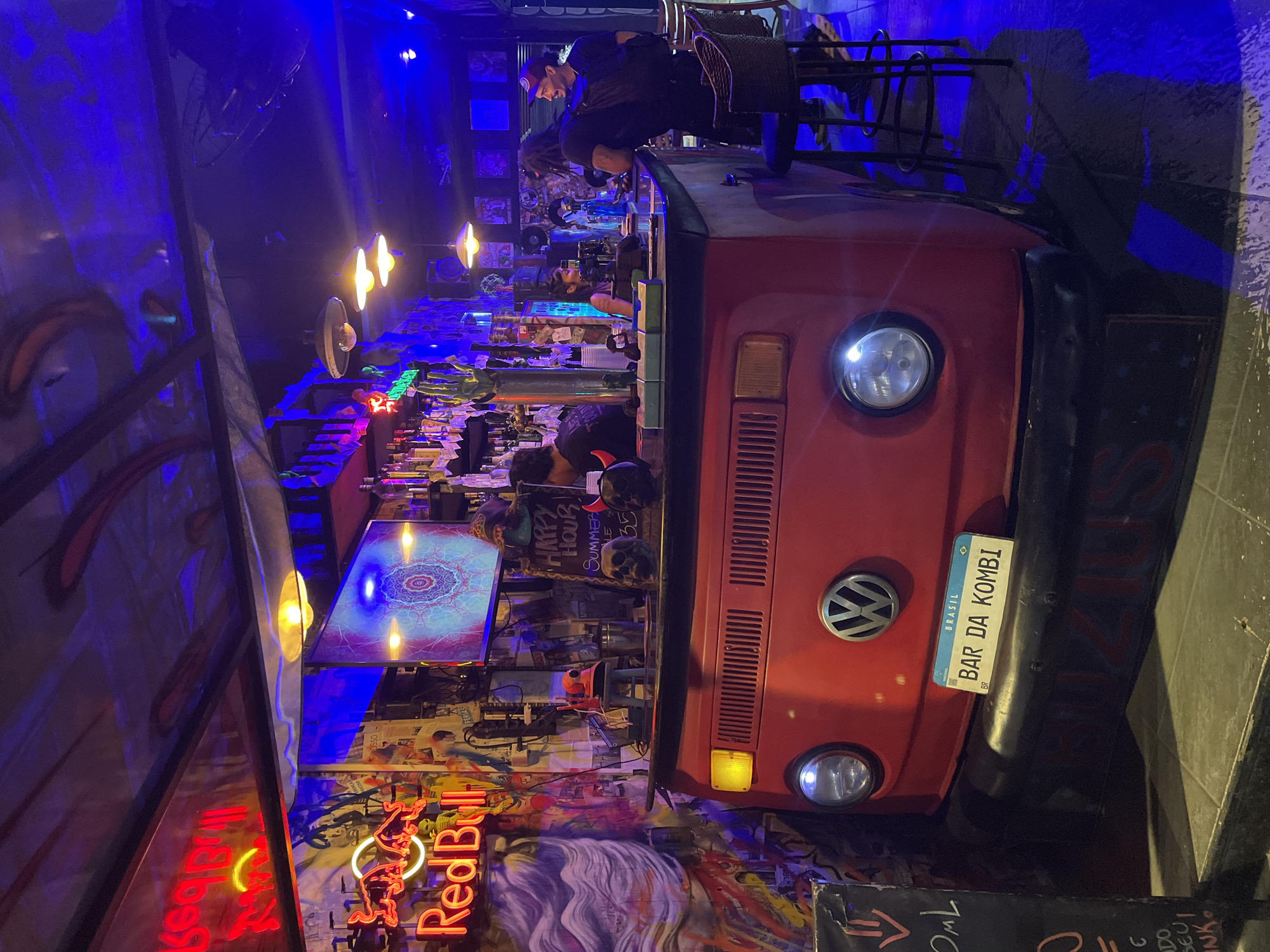 The image captures the interior of a bar in Armação Dos Búzios, Brazil, specifically "Bar da Kombi." The bar counter is made from a red Volkswagen Kombi van, with a license plate clearly visible. The bar area is brightly lit with a combination of neon signs (including one for "Red Bull"), spotlights, and illuminated display screens. Behind the bar, a person appears to be working. Additional people are seated or standing in the background. The walls are decorated with graffiti and various pieces of memorabilia, creating a vibrant and eclectic atmosphere.