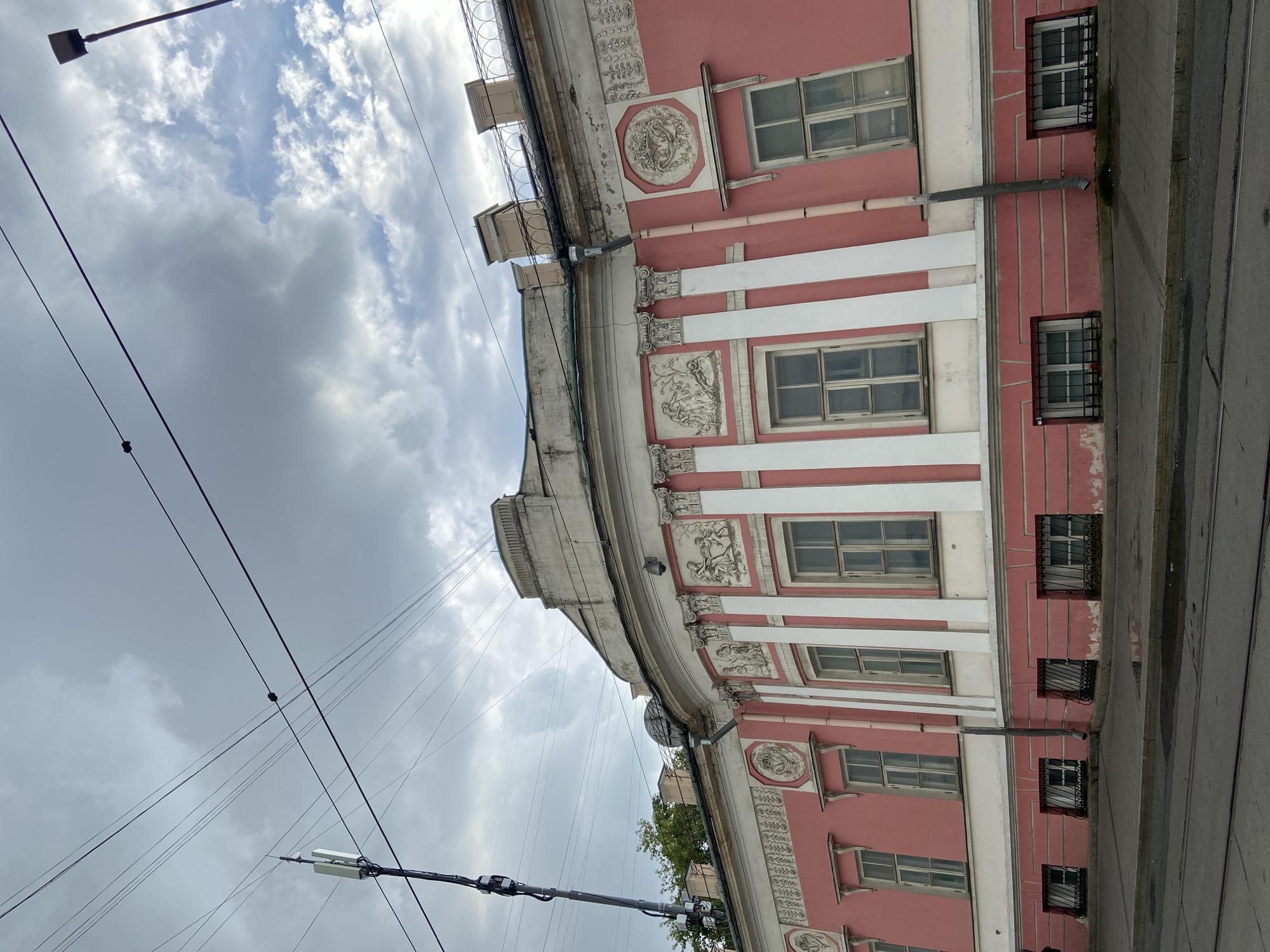 The image shows a curved, pink and white building with classical architectural elements, located in Moscow, Russia. The building has multiple windows on its facade, some with decorative bas-reliefs above them. There are also grilled windows on the lower level. In the foreground, a street or tram track is visible. Overhead, numerous power lines crisscross the sky, and a tall pole with what appears to be a cellular antenna is also present. The sky is cloudy, suggesting overcast weather. The building's architecture suggests it is an older structure, possibly a historical landmark.
