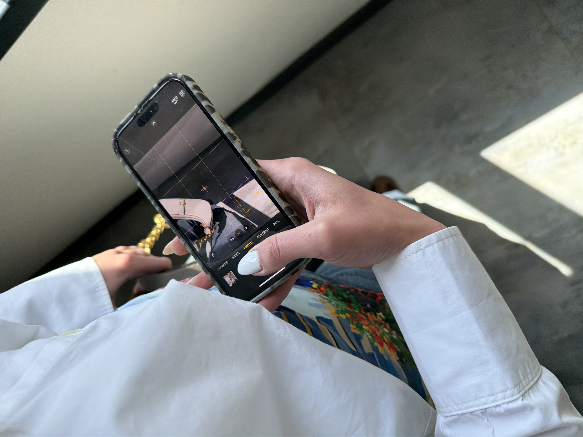 The image displays a close-up, overhead view of a person using a smartphone indoors in Yerevan, Armenia. The person's hands and forearms are visible, dressed in a white long-sleeved shirt with noticeable cuffs. A vibrant, colorful patterned fabric is partially seen beneath the white shirt, suggesting a skirt, dress, or another accessory.
The smartphone, an iPhone with a textured, possibly leopard print, protective case, is held in the person's hands. Its screen is active, showing the camera application interface. The camera mode is set to "PHOTO," with "CINEMATIC," "VIDEO," "PORTRAIT," and "PANO" modes also selectable. Zoom options "0.5x," "1x," and "2x" are displayed, with "1x" currently selected. The screen captures a partial view of a light-colored object, likely a handbag, featuring a distinctive golden chain. Grid lines and focusing aids, including a crosshair and a hash symbol, are visible on the display. Text at the top left of the screen reads "ƒNEg MAX," possibly indicating camera settings.
The setting appears to be a bright indoor space with a dark gray floor. A prominent rectangular area of direct sunlight falls across the floor, suggesting a nearby window and indicating a sunny daytime environment, likely during mid-day or afternoon. The background behind the person is softly blurred, appearing as a light gray or beige surface. The scene captures an individual in the act of photographing an item with their phone.