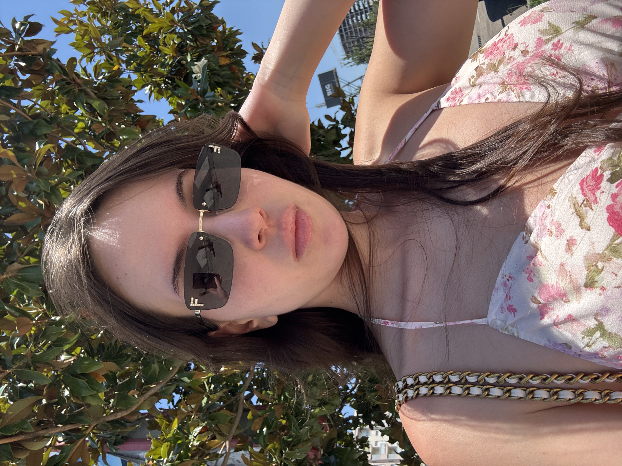A young woman is shown outdoors, lying down with her arm behind her head. She is wearing sunglasses with an "F" logo and a floral print top, with a gold and white chain accessory across her chest. Her hair is dark brown and cascades around her. She is surrounded by green leaves from a tree, and the sky is blue. The lighting suggests it is daytime, likely sunny. There is a building with the word "BIG" visible in the background. The overall impression is one of relaxation and leisure.