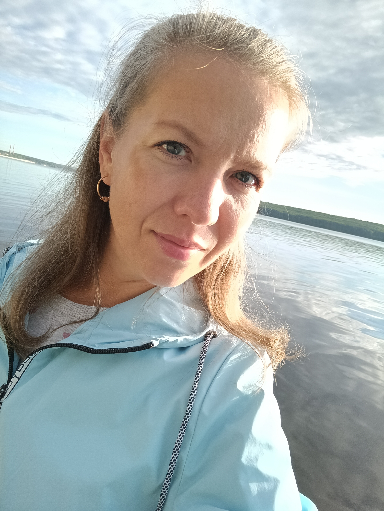 A woman with light brown hair and blue eyes is taking a selfie outdoors. She is wearing a light blue jacket over a grey shirt. She has a gold hoop earring in her left ear. The background shows a body of water, possibly a river, with a treeline on the far bank and a cloudy sky above. In the distance on the left bank, there are industrial structures, suggesting the image might be taken near Chaikovsky, Russia, which is known for its Kama River and industrial presence. The lighting suggests it is either early morning or late afternoon.