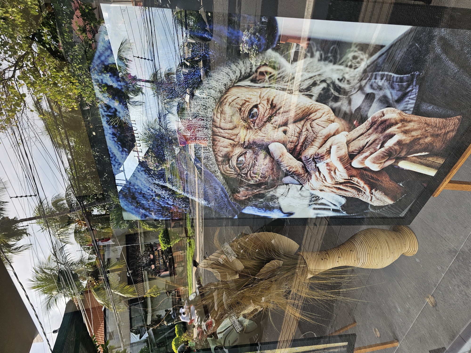The image, taken in Denpasar, Indonesia, shows a storefront window displaying a framed portrait of an elderly person with deeply wrinkled skin and wispy gray hair, smoking. The portrait is positioned above a decorative vase filled with dried plant material. The window reflects a street scene with palm trees, power lines, and buildings. Additional pedestrians and other objects are seen reflected in the window, making it difficult to definitively identify them. The scene is bright and taken during the day.