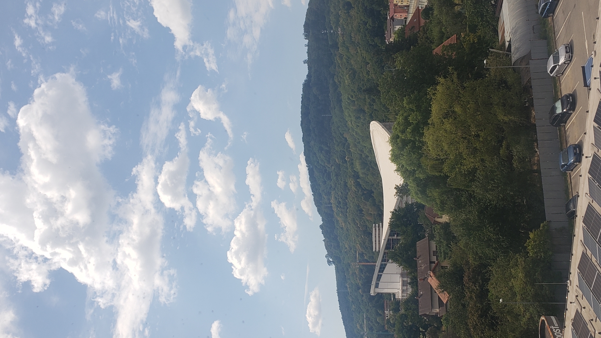 The image displays a scenic view of Prešov, Slovakia, dominated by a lush green hillside topped with a clear blue sky filled with white, puffy clouds. In the mid-ground, a large, modern building with a distinctive curved white roof is visible, possibly a stadium or arena under construction or renovation, indicated by scaffolding. Several smaller residential buildings with red-tiled roofs are nestled amongst the trees on the lower slopes of the hill. In the foreground, a parking lot with several cars, including a blue Audi, a dark hatchback, and a silver Audi, is visible. The parking lot appears to be adjacent to a commercial building with solar panels on its roof. The overall impression is one of a developing urban area surrounded by natural beauty.