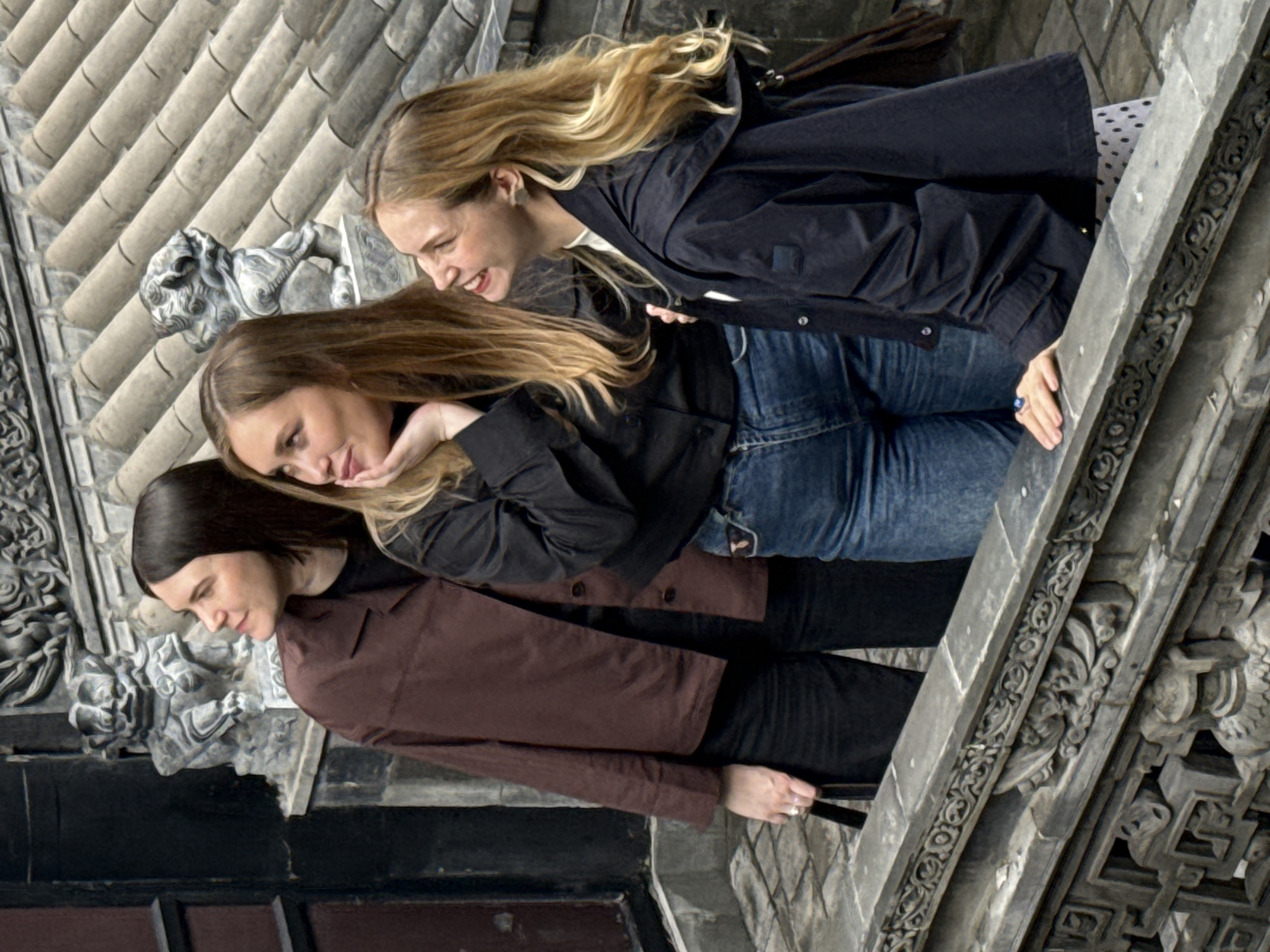 Three young women are depicted from the waist up, standing on what appears to be an ornate stone balcony or ledge, with intricate architectural details surrounding them. The image is oriented diagonally, but the women are upright within this frame.
The woman on the left (bottom in the original image) has dark hair and wears a dark brown jacket over dark clothing. She is looking downwards with a slight smile. The woman in the middle has long blonde hair, a black jacket, and blue jeans. She looks towards the woman on her left, resting her chin on her hand with a contemplative or amused expression. The woman on the right (top in the original image) also has long blonde hair and wears a dark jacket over a white top with black polka dots, and blue jeans. She is captured mid-laugh, with her head tilted back and a broad, joyful smile. Her right hand rests on the arm of the middle woman.
The background features a tiled roof with parallel ridges, and extensively carved stone elements, including a stone lion statue behind the women. The detailed carvings on the balustrade and the building suggest a traditional architectural style, consistent with the provided context of Yuzhong District, China. The scene takes place during the day, under natural light, and the women appear to be enjoying a moment of camaraderie and laughter. There is no visible text in the image.