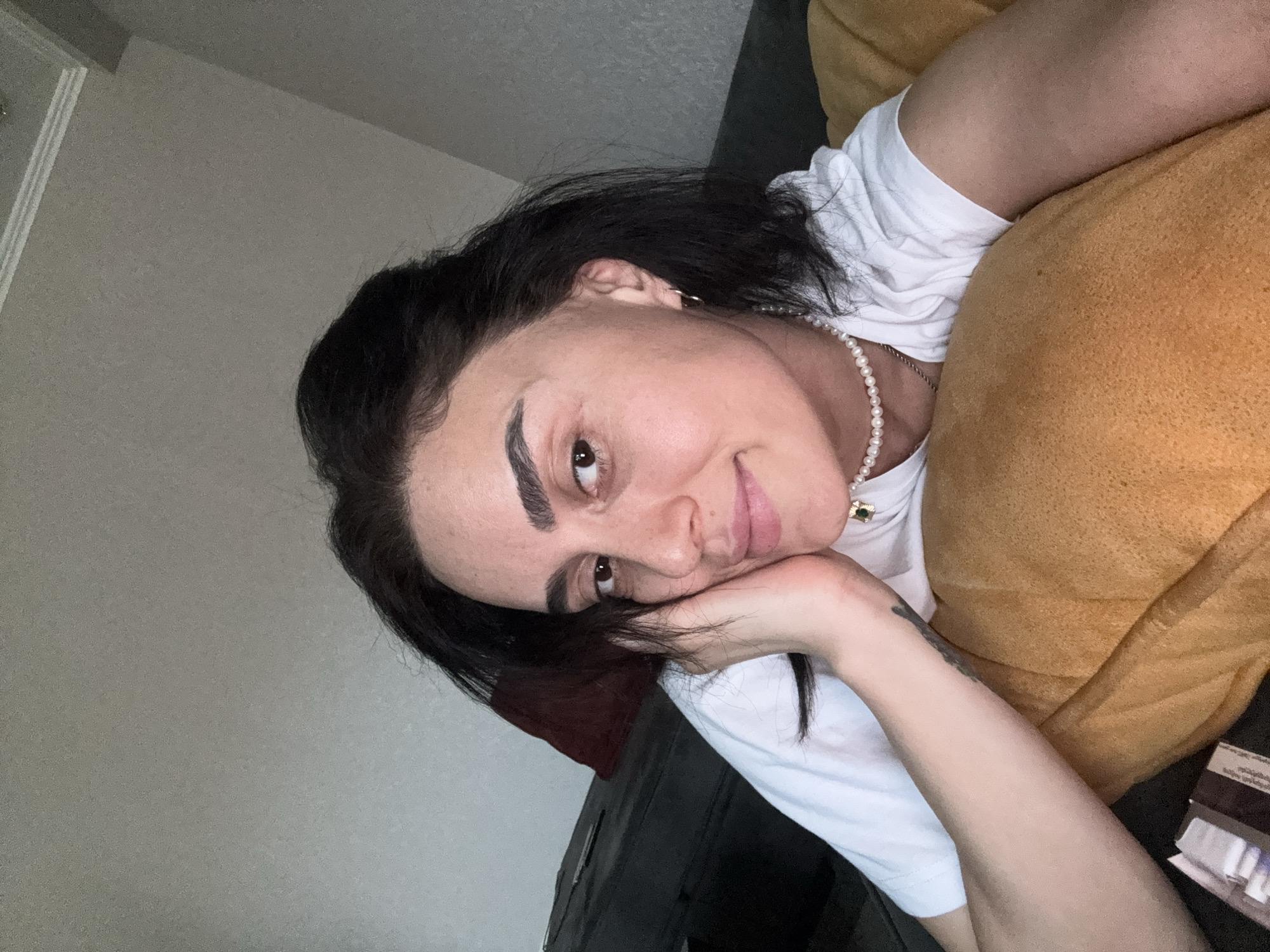 A woman with dark hair and brown eyes, wearing a white t-shirt and a pearl necklace, rests her head on a yellow pillow. She is lying on a couch in a room. The background shows a wall and a portion of the couch. There are no specific identifiable landmarks or features to indicate the city of Tbilisi, Georgia.