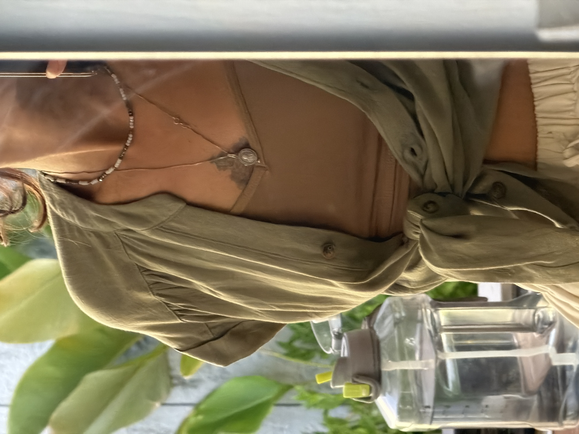 A medium shot shows a person from the chest down, wearing a light brown tank top and an olive green unbuttoned shirt tied at the waist. The person is also wearing light beige pants. Two necklaces are visible, one with small beads and another with a circular pendant. Behind the person, large green leaves are visible. To the left, a large clear plastic water jug with a grey and yellow lid and a white straw is visible. The scene appears to be outdoors, possibly on a balcony or patio, with a hint of wooden siding in the background. The lighting suggests it is daytime. The scene is set in the city of Kuta Selatan, Indonesia.