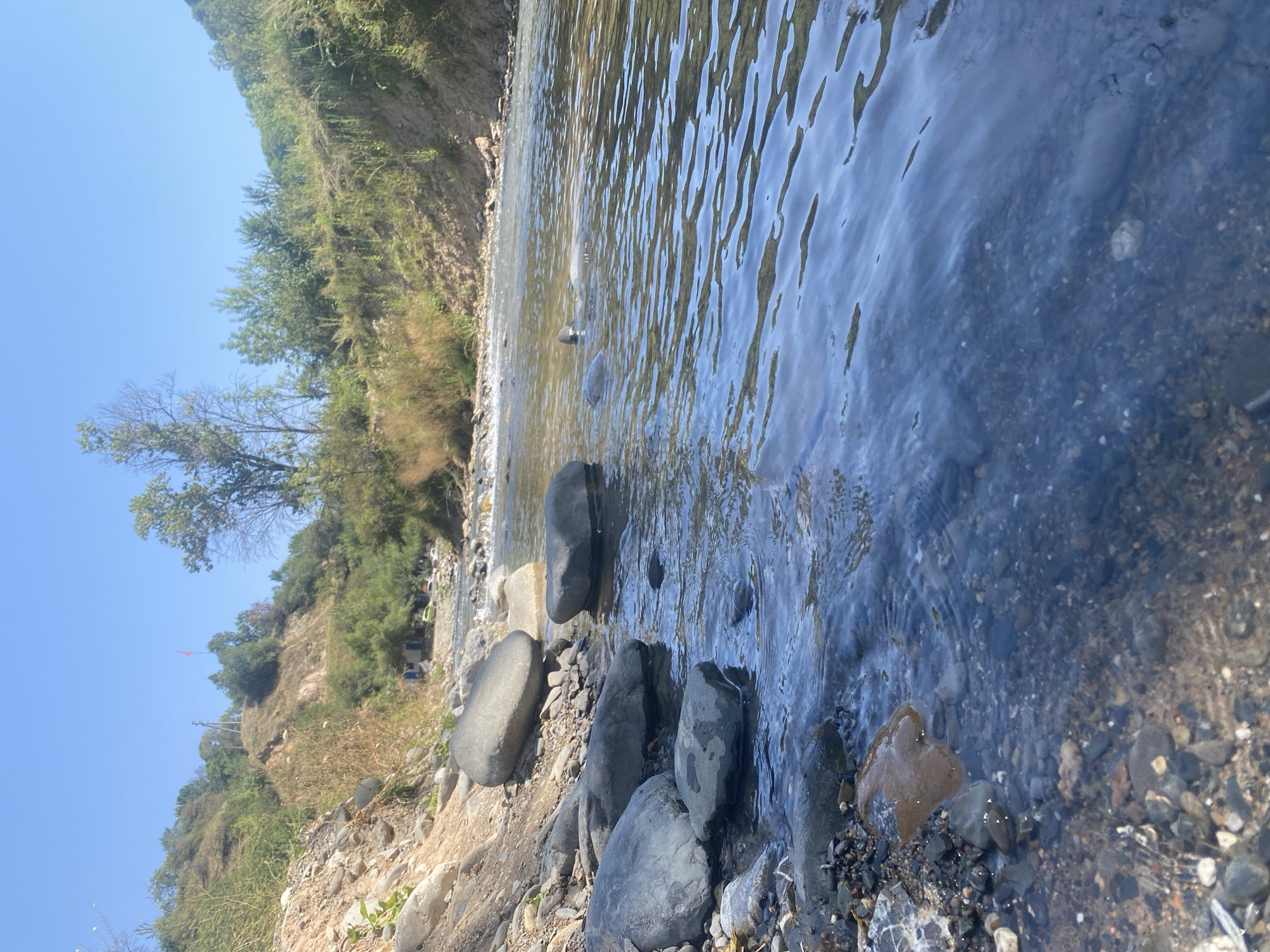 The image shows a natural landscape with a clear blue sky and a river flowing through a valley. The foreground is dominated by a rocky riverbank with several large, smooth stones partially submerged in the water. The river itself is shallow and appears to be flowing over a bed of smaller pebbles and rocks. The water surface is rippled, reflecting the sunlight and the surrounding greenery.
The background consists of steep, grassy hillsides covered in vegetation, including trees and bushes. In the distance, along the riverbank, there are some small structures that appear to be houses or sheds, with a few vehicles parked nearby. A red flag is visible on top of one of the hills. The overall impression is one of a peaceful, rural setting in a mountainous region. The location is identified as Kara-Alma, Kyrgyzstan.