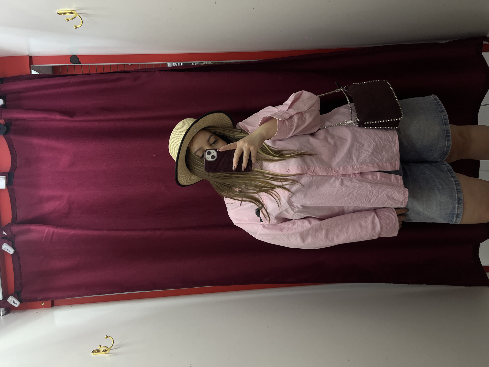 A person is taking a selfie in what appears to be a dressing room. They are wearing a straw hat, a light pink button-down shirt, and denim shorts. A small brown purse is slung over their shoulder. The background consists of a maroon curtain and white walls. There is a hook visible near the top left of the frame. There is no indication of the location being Novopokrovskaya, Russia.