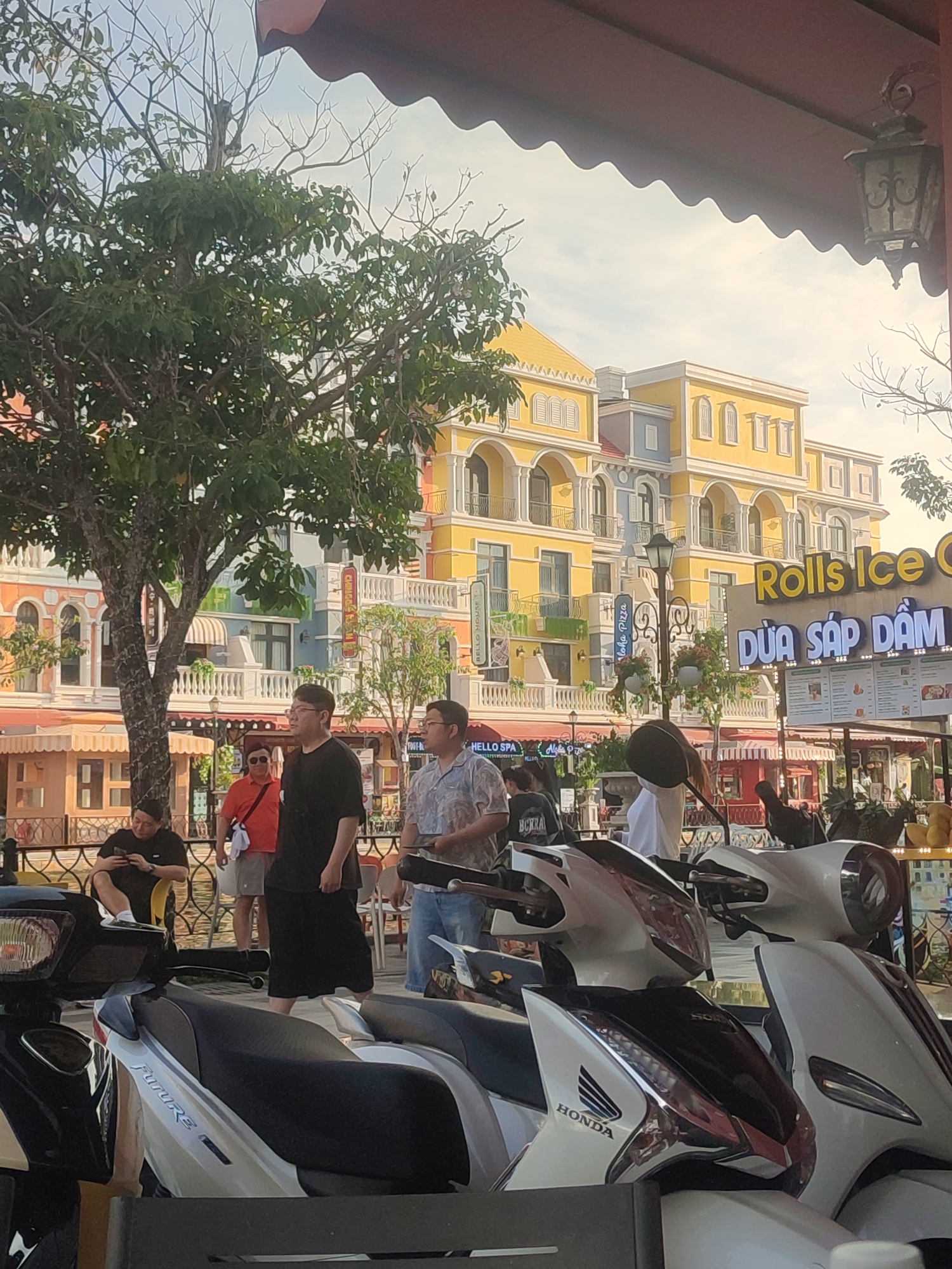 The photograph captures a street scene in Phú Quốc Special Administrative Region, Vietnam. In the foreground, several white and black Honda Future scooters are parked, partially obstructing the view. Behind the scooters, a group of people is seen. Among them, a man in a black t-shirt and black shorts stands out, along with another man in a patterned shirt and jeans. Further back, a woman in a red shirt and shorts walks by. A man sitting on a bench is looking at a phone.
The backdrop features colorful buildings with a distinct European architectural style, painted in hues of yellow, orange, and blue. Signage for "Rolls Ice Cream" and "Dừa Sáp Dằm" is visible, indicating the presence of a dessert establishment. There is also signage for 'HELLO SPA' and 'Aloha Pizza'. A large tree with green foliage stands on the left side of the frame. The photo is taken from under an awning, which is visible at the top of the image.