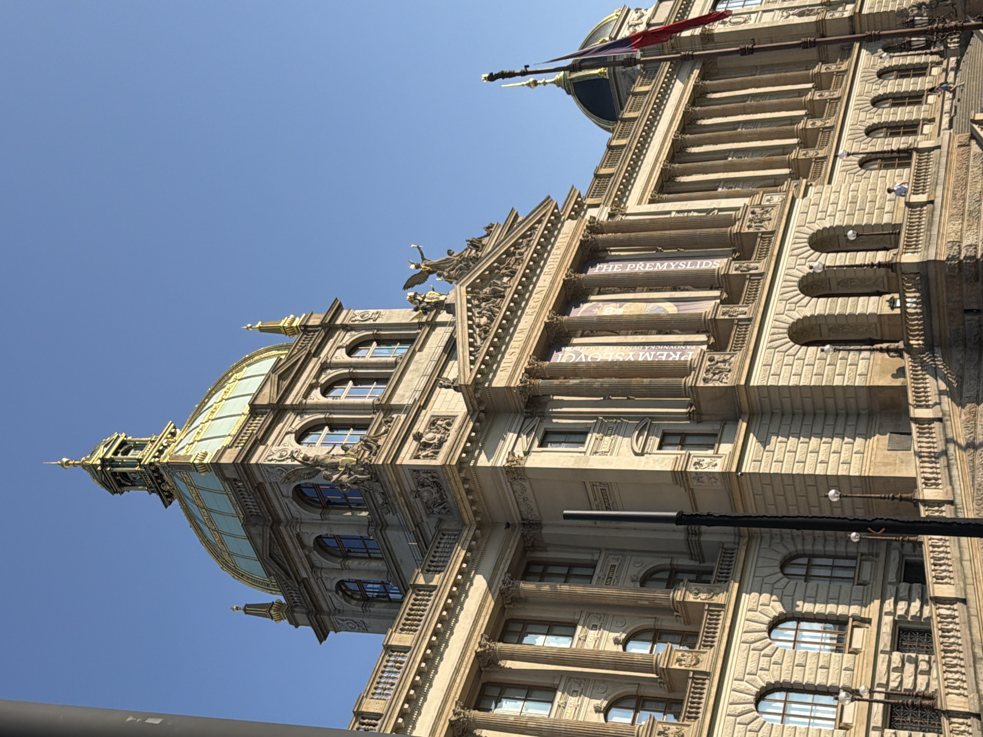 The media file captures an upward-angle view of a grand, ornate building, likely in Prague, Czechia, against a clear blue sky, suggesting a sunny day. The building itself is a prominent subject, characterized by its intricate architectural details, including classical columns, arched windows, decorative carvings, and a distinctive dome topped with a spire. The facade is constructed from stone, giving it a historical and imposing appearance. Several banners are visible on the building's exterior, displaying text. One banner reads "THE PREMYSLIDS," and another partially visible banner appears to be in a language other than English, possibly Czech. The building's design suggests it might be a museum or a significant public institution. The perspective from below emphasizes the scale and height of the structure. There are no people clearly visible in the frame, but the presence of flags, though partially obscured, hints at a possible event or national symbolism associated with the location. The lighting and shadows suggest it is daytime, likely midday, given the bright illumination. The overall impression is one of historical grandeur and architectural magnificence.