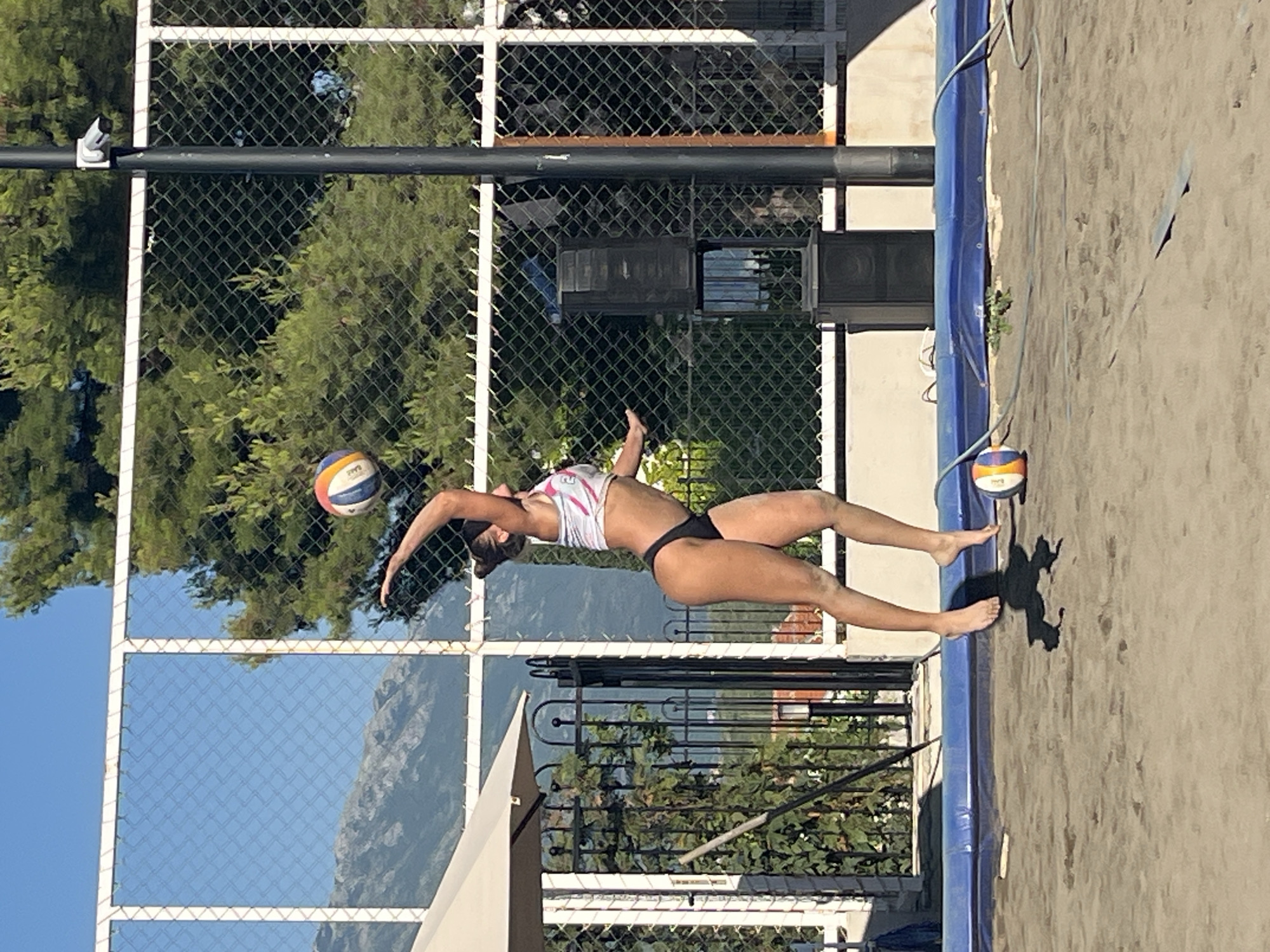 In Gošići, Montenegro, a woman is captured mid-air on a sandy beach volleyball court, executing a volleyball serve. She is wearing a white and pink athletic top and black bikini bottoms. Her body is positioned in a dynamic pose, with her right arm extended upwards, about to strike the volleyball. The volleyball is in the air above her head, with blue, orange, and white panels visible. Her left arm is extended downwards for balance, and her legs are bent as she is in the process of jumping. Her shadow is cast on the sand below.
In the background, a chain-link fence with a white top rail encloses the court. Beyond the fence, lush green trees and a mountain range under a clear blue sky are visible. A black volleyball pole stands to the right of the frame, with a security camera mounted near the top. Two black speaker units are also situated behind the fence. A large umbrella and part of a fence are visible to the left of the court. Another volleyball lies on the sand to the right of the woman. The lighting suggests it is daytime with the sun casting shadows.