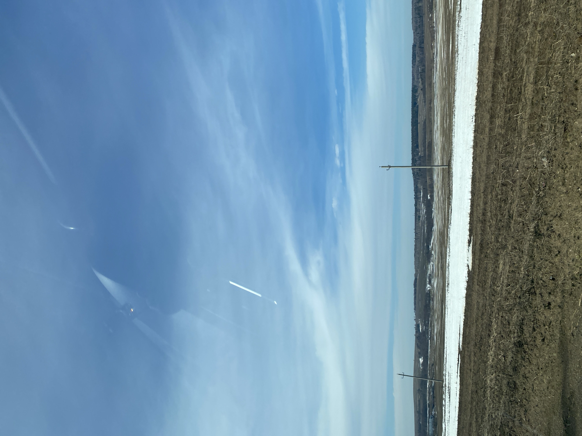 The image depicts a rural landscape on a sunny day, likely in the vicinity of Буль-Кайпаново, Russia given the prompt. A clear blue sky with scattered clouds occupies the left portion of the image. The right side of the frame shows a field, with areas of snow cover interspersed between patches of brown soil. Two utility poles are visible, sparsely spaced along the border between the field and the sky. Some trees are visible near the horizon line. Reflections are seen in the glass the photograph was taken through, adding artifacts.