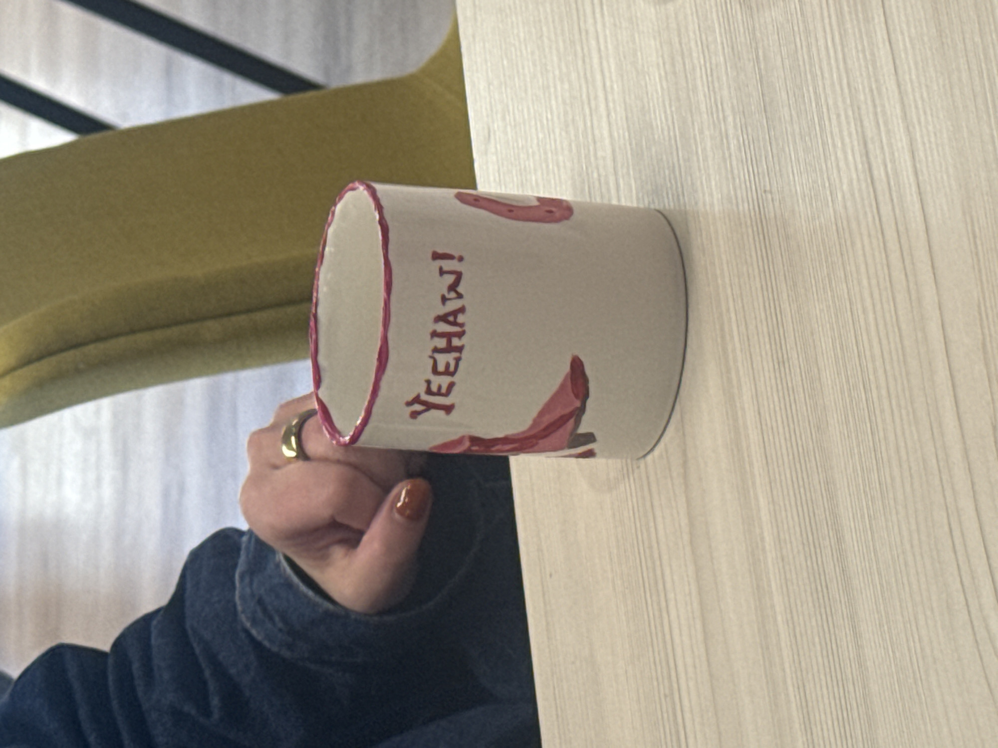 A hand wearing a gold ring and with a brown-colored fingernail on one finger holds a white cylindrical mug with a pink-trimmed edge. The mug has the word "YEEHAW!" written in red and has red images of a cowboy boot and horseshoe. The mug rests on a light-colored wooden surface with a linear pattern. A portion of a yellow chair is visible in the background. The person holding the mug is wearing a dark blue denim jacket. The photograph appears to be taken indoors.