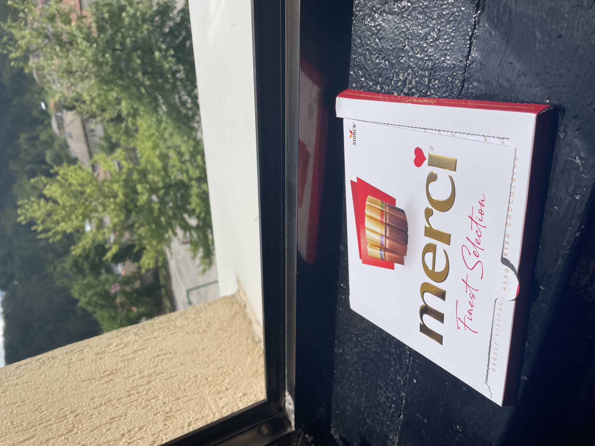 A box of Merci Finest Selection chocolates is lying on a black, textured surface. The packaging is white with gold and red accents. The chocolates are partially visible on the box. The box appears to be positioned near a window frame with a beige interior and black exterior. Outside the window, greenery and buildings are visible, suggesting an outdoor urban or suburban environment. The image was taken in Zlatograd, Bulgaria.