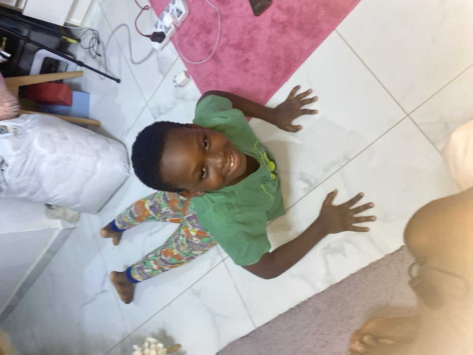 A smiling boy lies on the floor, posing like a star. He wears a green shirt and patterned leggings. His hands are outstretched, touching the white tiled floor. Behind him are a pink rug and a white object. The photo appears to have been taken from above and the picture appears to be rotated 90 degrees.
