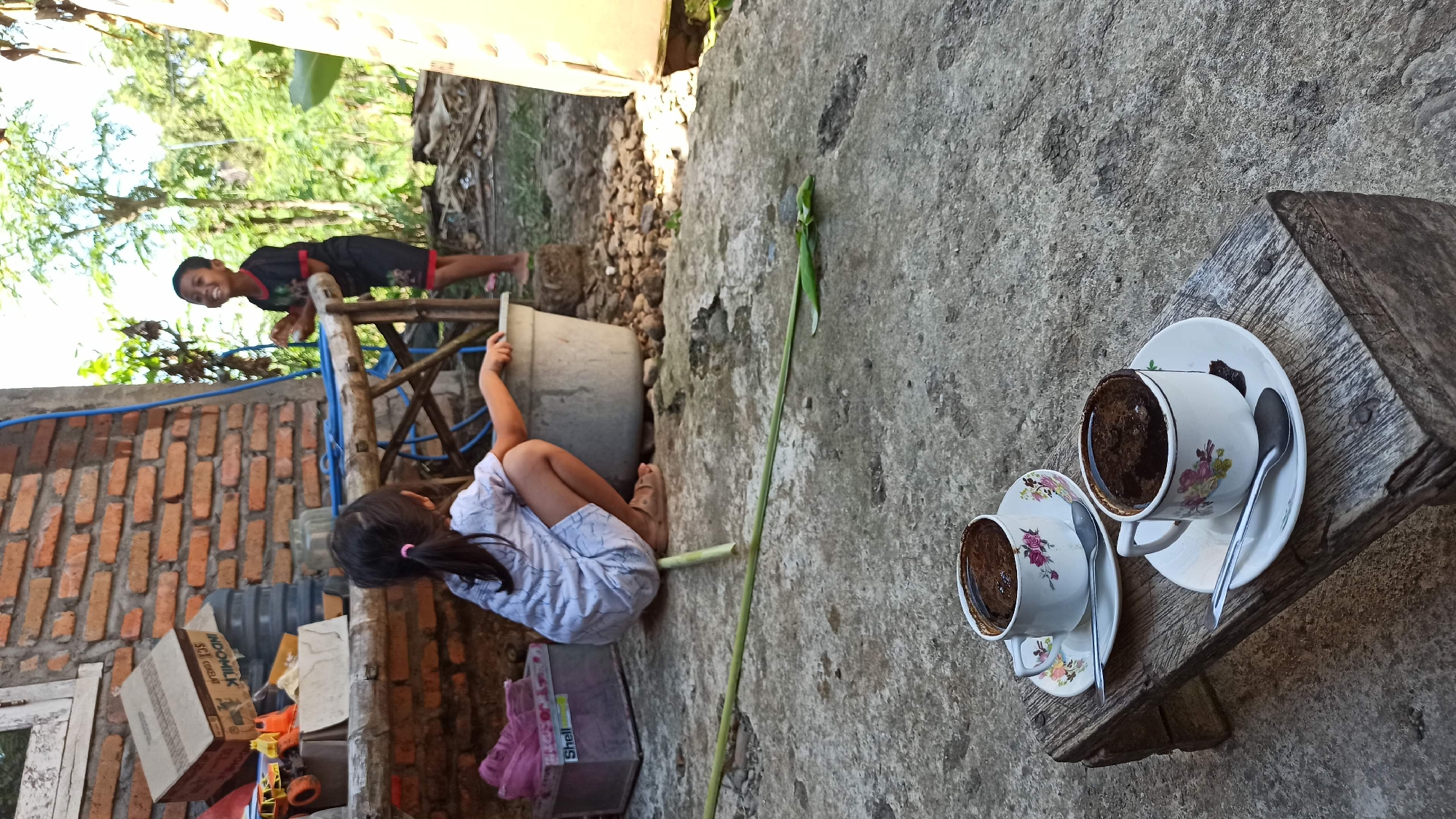 In Gegeran, Indonesia, two children, a girl and a boy, are engaged in outdoor activities. The girl, wearing a white shirt and shorts, is crouching by a large grey basin, her right hand near its rim. She has a ponytail with a pink tie. The boy, in a black t-shirt with red trim and black shorts, stands behind a wooden table, laughing. The scene takes place on a rough, concrete ground. In the foreground, two cups of dark coffee are placed on a rustic wooden stool. Each cup sits on a saucer with a floral pattern and is accompanied by a spoon. Behind the children, a brick wall stands partially weathered. Scattered items, including toys, a cardboard box labeled "INDOMILK," and a purple container, are visible on and around the wooden table. A green plant stem lies horizontally on the concrete. The background features lush green trees, indicating a rural or semi-rural setting.