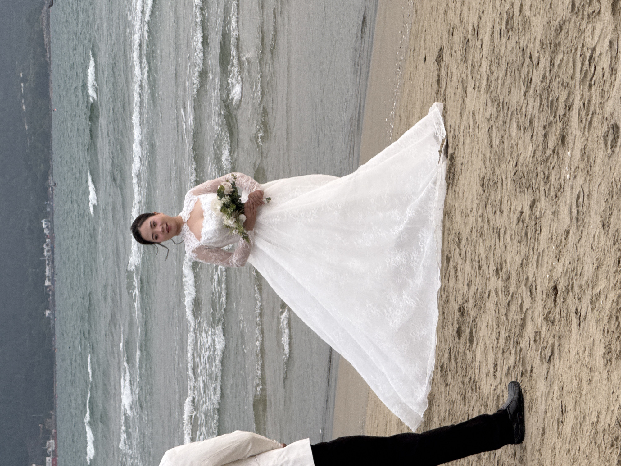 A bride in a white lace wedding dress and holding a bouquet stands on a beach in Đà Nẵng, Vietnam. Her dress trails onto the sandy beach. The ocean with visible waves is adjacent to the beach. Partially visible at the bottom of the frame is a person wearing black pants, a black shoe, and a white shirt. A city can be seen in the distance along the coastline.