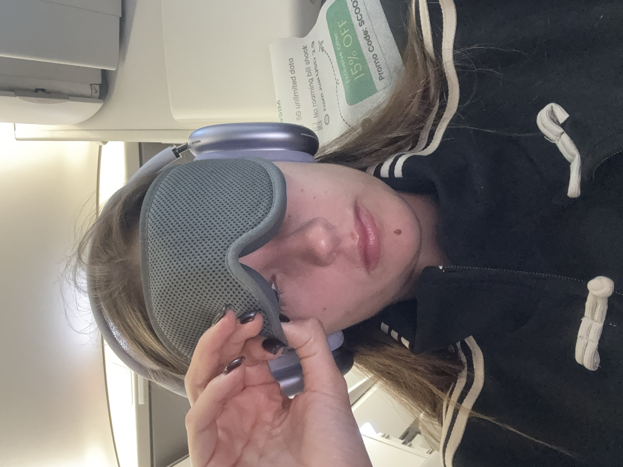 A young woman is pictured inside an airplane cabin. She is wearing a gray eye mask and gray headphones, and is holding the edge of the eye mask with her left hand. She is wearing a black jacket with white stripes down the arm. A paper advertisement with a "15% OFF" promotion code is positioned near her head. The text on the advertisement mentions "5G unlimited data" and "No roaming bill shock." The location is identified as being in ตำบล ไม้ขาว, Thailand.