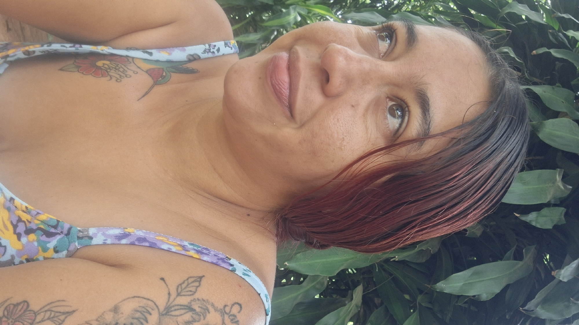 This close-up portrait, taken outdoors in Vallecito, Venezuela, features a woman looking upwards and slightly to the viewer's right. Her face is well-lit by natural daylight, suggesting a clear daytime setting. She has medium-toned skin and her dark hair, with prominent reddish highlights, falls across her face and shoulder, appearing somewhat wet or sleek. Her brown eyes are open, reflecting the light, and her lips are gently parted in a subtle, contemplative smile.
She is wearing a light-colored top with a patterned design in shades of light blue, purple, yellow, and green. Multiple tattoos are visible on her upper body. On her upper left chest, near her collarbone, a colorful tattoo depicts a green and red hummingbird feeding from a red flower with yellow details. On her right shoulder and upper arm, outlines of floral or vine-like tattoos are visible, partially obscured, and may include an animal figure or text that is not clearly legible.
The background is dominated by dense, lush green foliage with large leaves, indicating a natural, possibly tropical or subtropical, outdoor setting like a garden or forest. The overall scene conveys a calm and serene atmosphere.