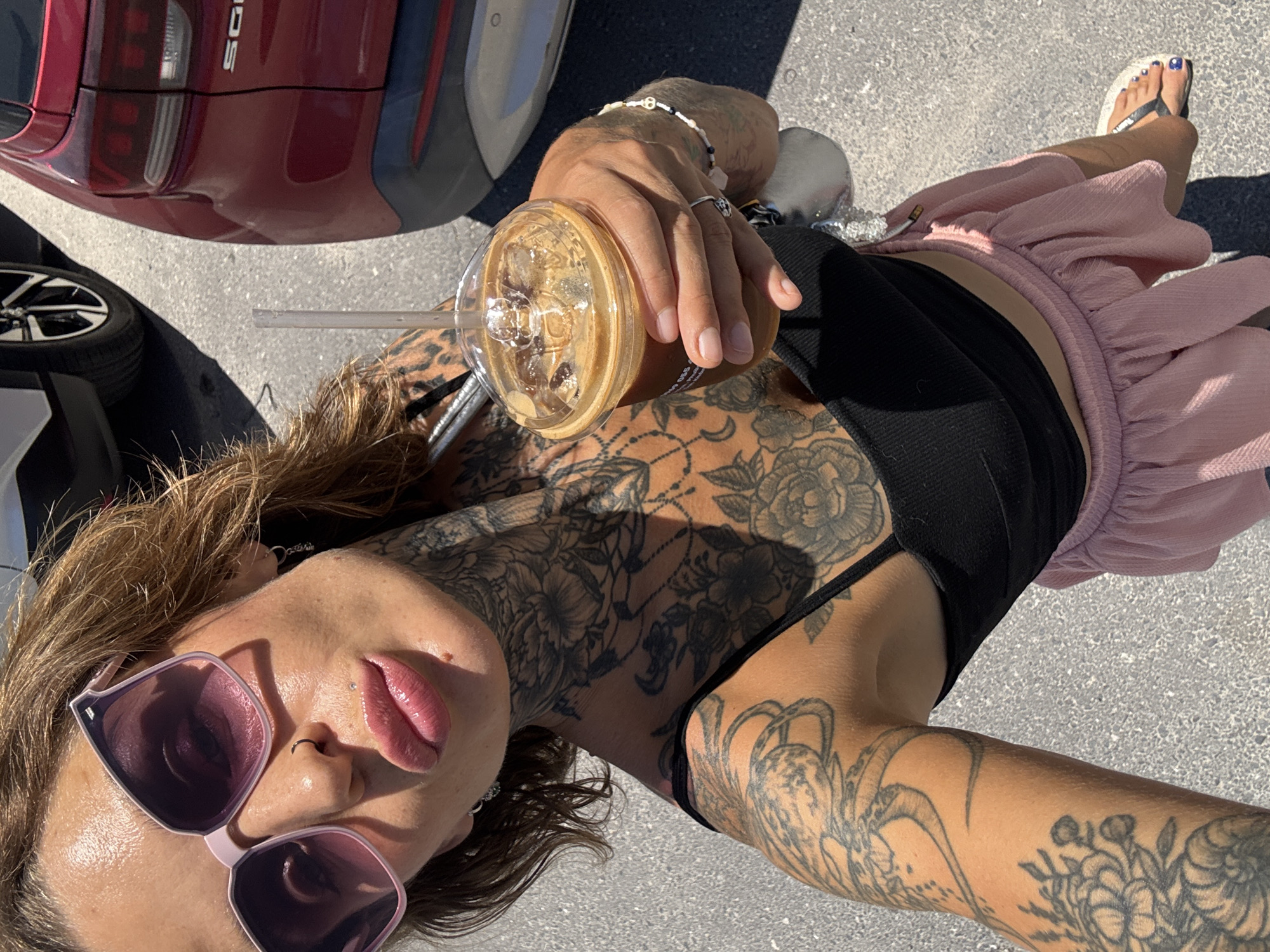 A woman with extensive tattoos on her arms, neck, and chest takes a selfie while holding a cold beverage. She is wearing large pink sunglasses, a black crop top, and a pink ruffled skirt. Her lips are highlighted with lip gloss. She is standing outdoors on a paved surface, with a red car visible in the background.