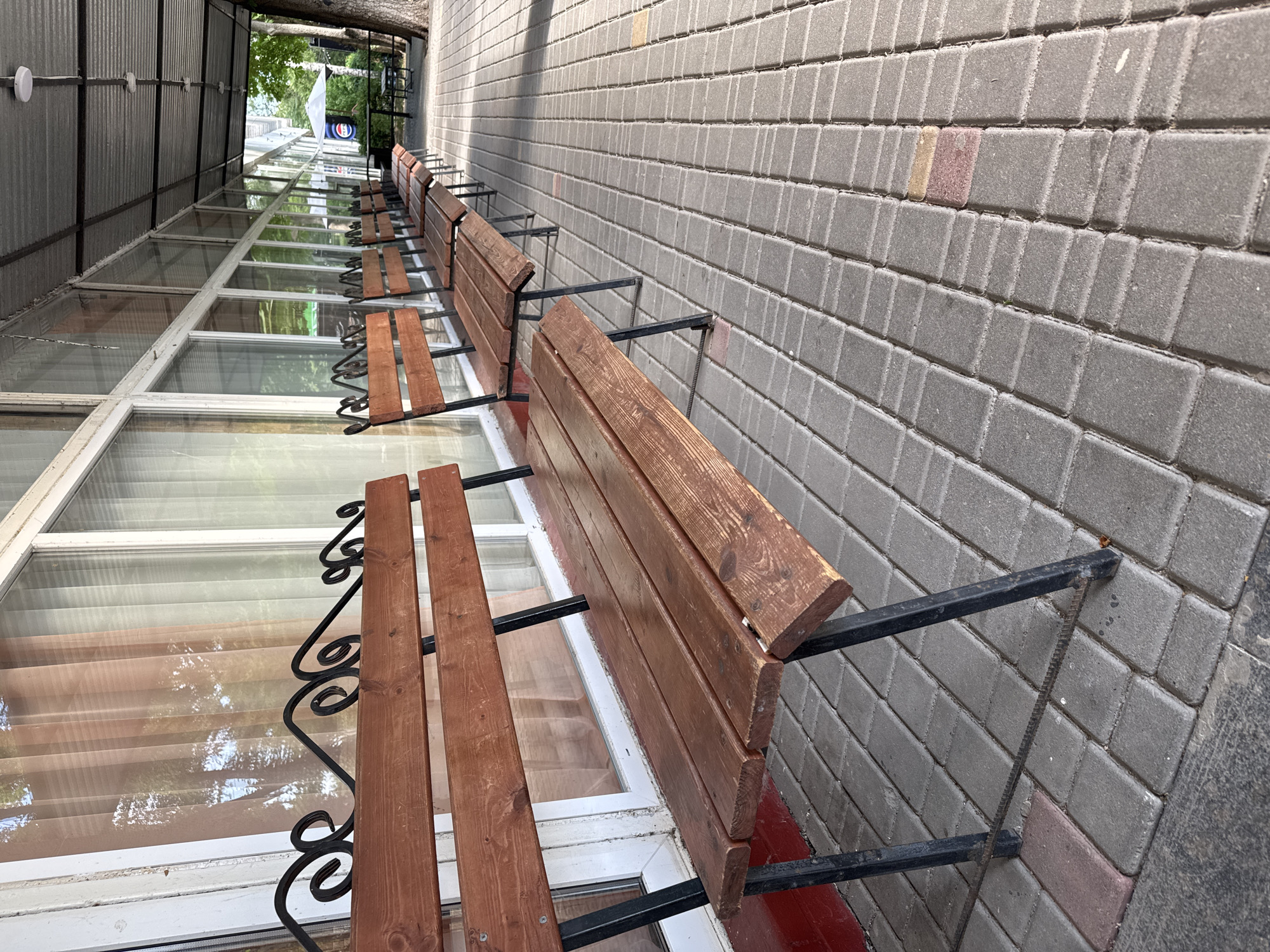 The image shows a row of wooden benches attached to a brick wall. The benches are made of dark red wooden planks and are supported by black metal frames. They are mounted on a paved walkway, and behind them is a building with large windows. The scene is outdoors, under an overhang, suggesting a covered outdoor seating area. The lighting indicates it is daytime, and the weather appears to be clear. The building's architecture and the style of the benches suggest a location in a public or commercial space, possibly a cafe or restaurant. The wall is made of grey interlocking pavers. Some of the pavers are lighter in color, possibly due to weathering or different materials. There is a red base visible at the bottom of the wall. The windows reflect trees and the sky, indicating greenery in the surroundings. The image is taken from a slightly elevated angle, looking down the row of benches. No people are visible in the image. There is no visible text in the image.