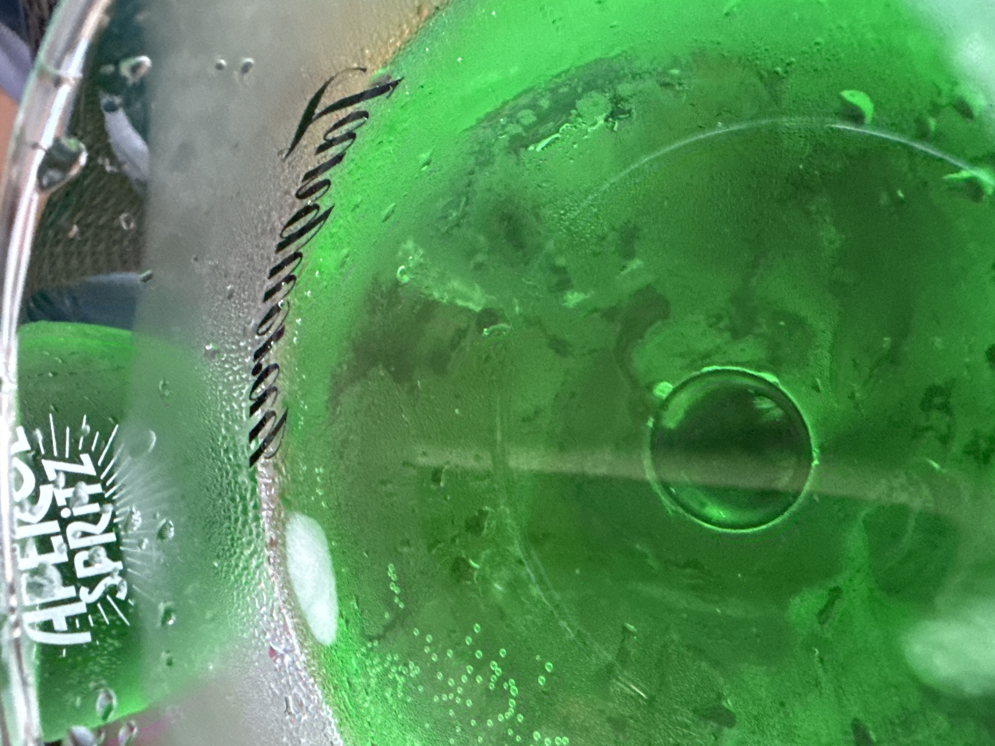 This media file is a close-up, top-down shot of a glass containing a green beverage. The glass is covered in condensation, with visible water droplets and streaks. Within the drink, there are numerous small bubbles.
On the exterior of the glass, there is white text that appears to be a logo or branding. The visible portion reads "APEROL SPRITZ," with "APEROL" in a stylized font with radiating lines above it, and "SPRITZ" below. There is also some black script text on the condensation-covered part of the glass, which is difficult to decipher completely but appears to be a signature or decorative element.
The setting appears to be outdoors, possibly on a warm day given the condensation. The color of the drink suggests it might be an alcoholic beverage like Aperol Spritz. While no people are directly visible, the context of a drink being consumed implies human presence. The image's focus is entirely on the drink and its container. The lighting suggests it could be daytime. The context provided places this scene in Sozopol, Bulgaria.