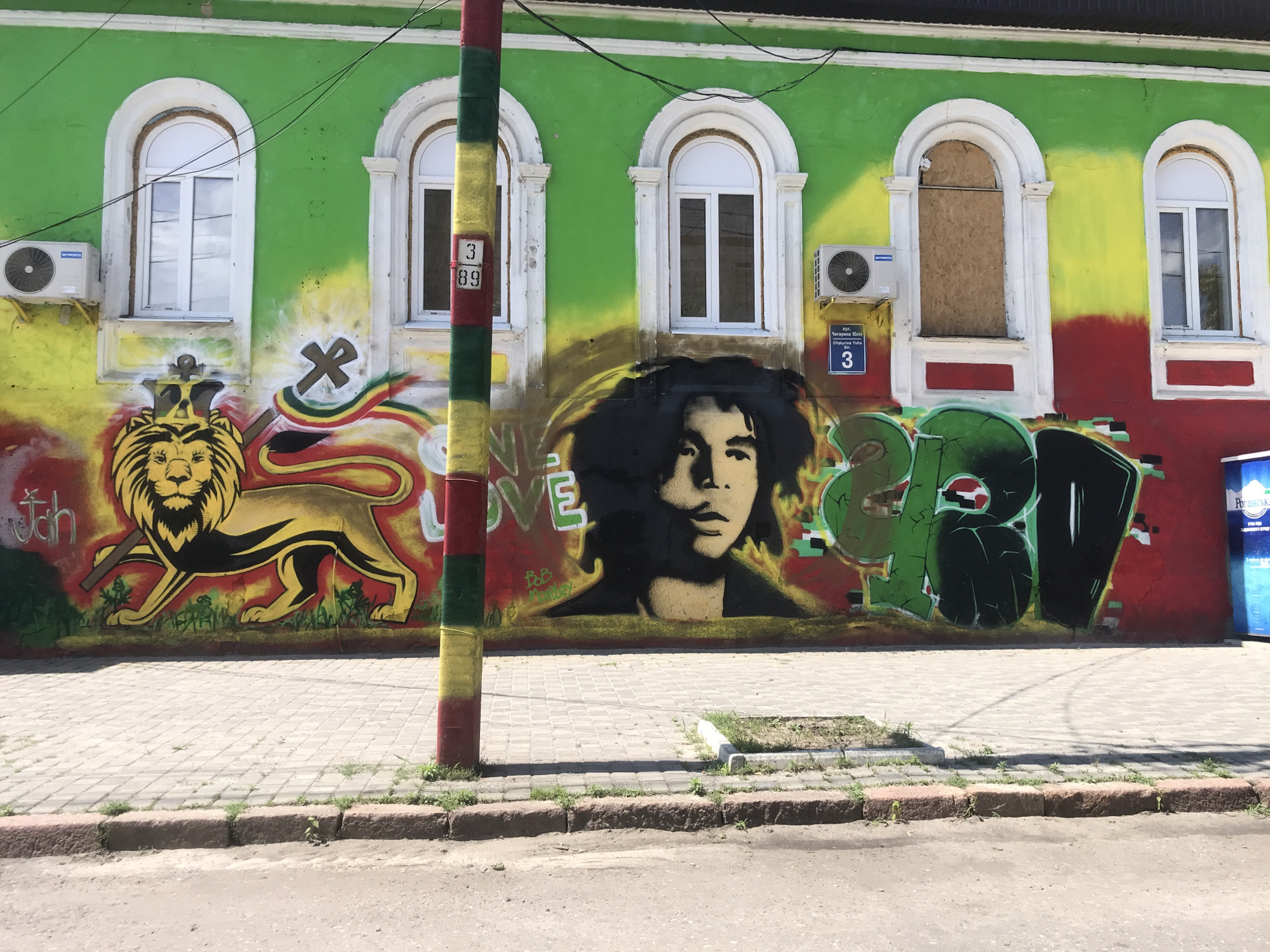 The image shows a vibrant mural on the exterior wall of a building in Kharkiv, Ukraine. The mural features a large, stylized depiction of Bob Marley with his signature dreadlocks, positioned centrally. To the left of Marley's portrait is a roaring lion with a crown, symbolizing Rastafarianism, alongside the word "Jah." To the right of Marley's portrait, large, graffiti-style lettering in green and black spells out the word "REVOLUTION." The building itself is painted with broad stripes of green, yellow, and red, colors strongly associated with Rastafarian culture. There are three arched windows on the building's facade, two of which are white-paned, and one is boarded up. A striped pole, painted in the same green, yellow, and red, stands in front of the mural, partially obscuring the left side of the artwork. The building's wall also displays two air conditioning units and various street signs and numbers, including a blue street sign with Cyrillic text and the number "3." A paved sidewalk runs along the base of the wall, with a street visible in the foreground. The scene appears to be a public space, likely in a residential or commercial area of the city, given the presence of windows and street infrastructure.