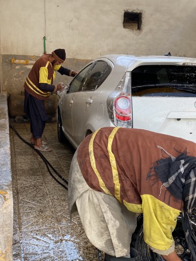 Two men are washing a white car in an enclosed space. The man on the left is wearing a brown and yellow sweater, dark pants, and a dark cap, and is actively washing the side of the car. The other man, closer to the camera, is bent over, partially obscuring the view, and is wearing a brown and yellow shirt. The floor is wet and covered in soap suds, and there is a black hose on the ground. The walls are unfinished concrete. The scene appears to be taking place in a car wash facility, but specific details confirming its location in Kandahar, Afghanistan are not evident in the image.