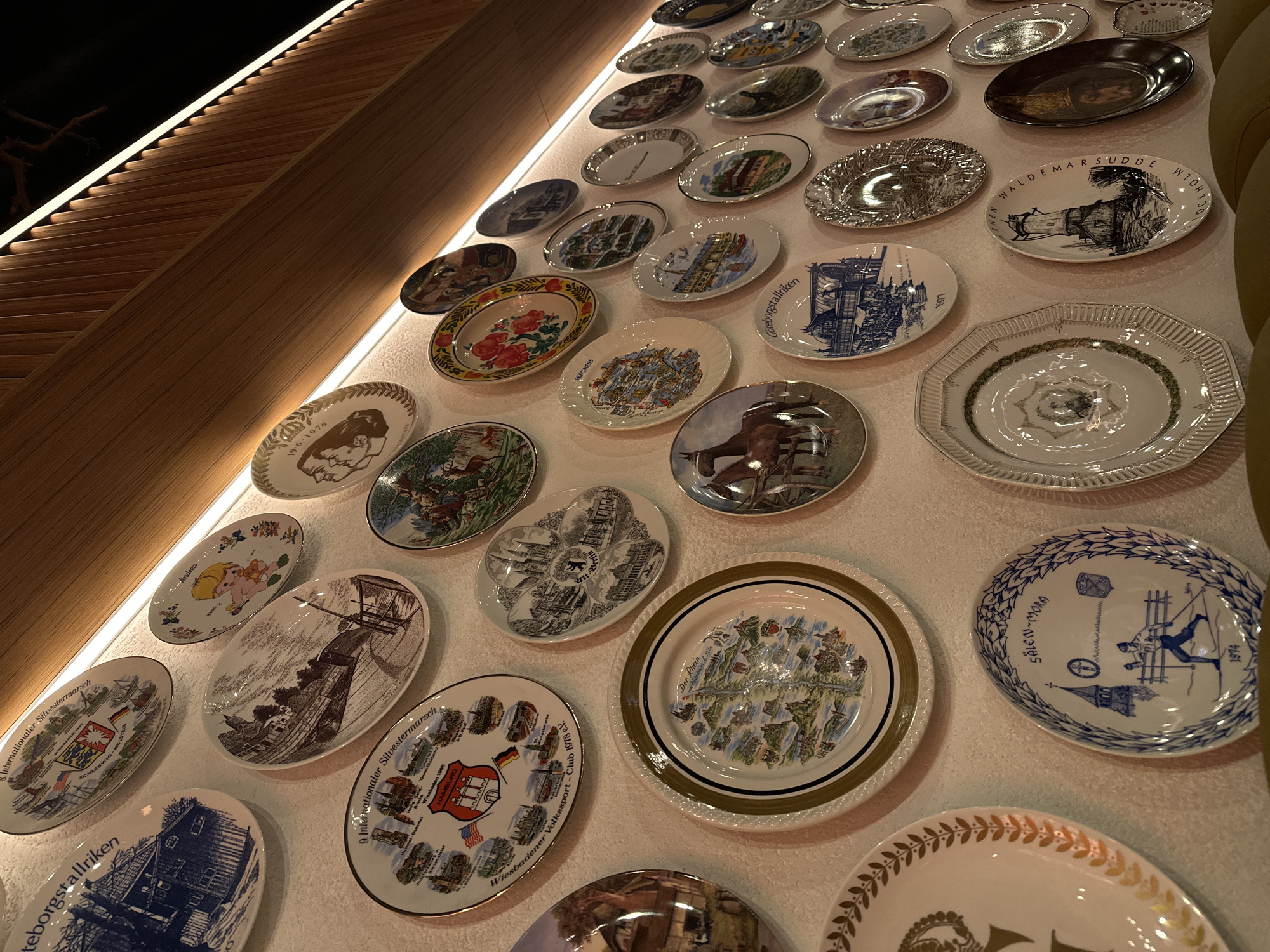 The image shows a wall decorated with a collection of decorative plates. The plates vary in design, color, and size, and are arranged in rows. Some plates are white with blue or black illustrations, while others have colorful floral or landscape designs. The plates are mounted on a white wall, which is illuminated by a linear light source. The plates appear to be arranged in a somewhat organized manner, possibly as part of a display or art installation. The plates also display names of places.