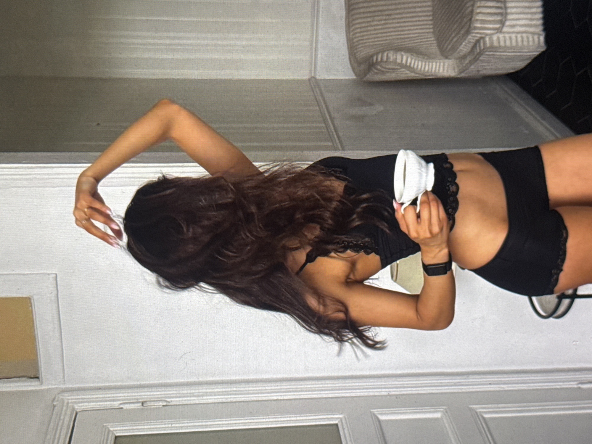 A person with long, dark hair is holding a white mug and appears to be indoors. They are wearing black undergarments. The person is positioned in the center of the frame, with their body turned away from the camera. Their right arm is bent, holding the mug, and their left arm is also bent, with their hand near their head. The background consists of a white wall and a door, suggesting an interior setting. The lighting appears to be natural, possibly from a window, and there are no strong shadows, indicating it might be daytime. The image has a casual and relaxed feel. There is no visible text in the image.