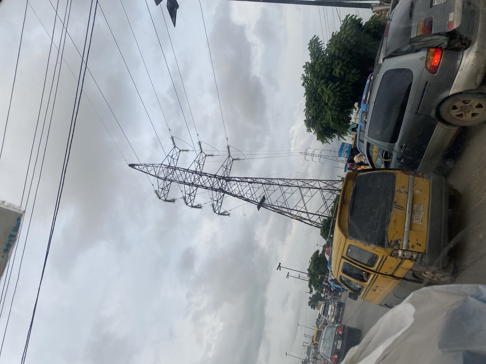 The image depicts a traffic scene in Ikeja, Nigeria, under an overcast sky. A tall, lattice metal electricity pylon dominates the right side of the frame, leaning slightly. Several power lines crisscross the sky above the scene. In the foreground, a yellow and grey minibus, appearing worn, is stationary in traffic. To its right, a grey minivan is also stopped. Other vehicles, including a dark sedan, are visible further back in the congested roadway. Pedestrians are seen on the side of the road, with some gathering near the vehicles. Lush green trees are situated in the middle ground. The overall atmosphere suggests a typical urban traffic situation in Ikeja, possibly during rush hour or due to an obstruction.