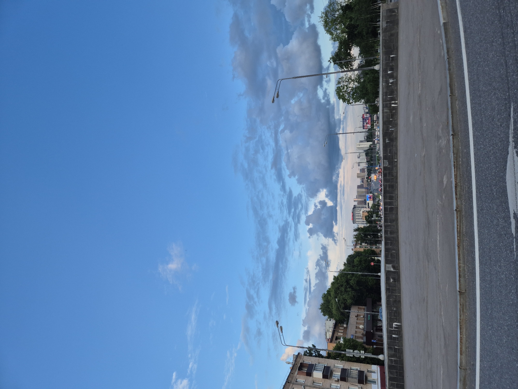 This is a ground-level, eye-level, medium shot of a street scene in Moscow, Russia. The sky is a bright, clear blue with scattered white clouds. In the foreground, there is a grey asphalt road with a white line marking. Behind the road, there is a grey concrete barrier with a dark tiled wall. To the left of the barrier, there is a multi-story building with balconies. In the background, there are trees, streetlights, and a city skyline with tall buildings. There are also some cars and billboards visible. The overall scene suggests a cityscape with urban infrastructure and natural elements.