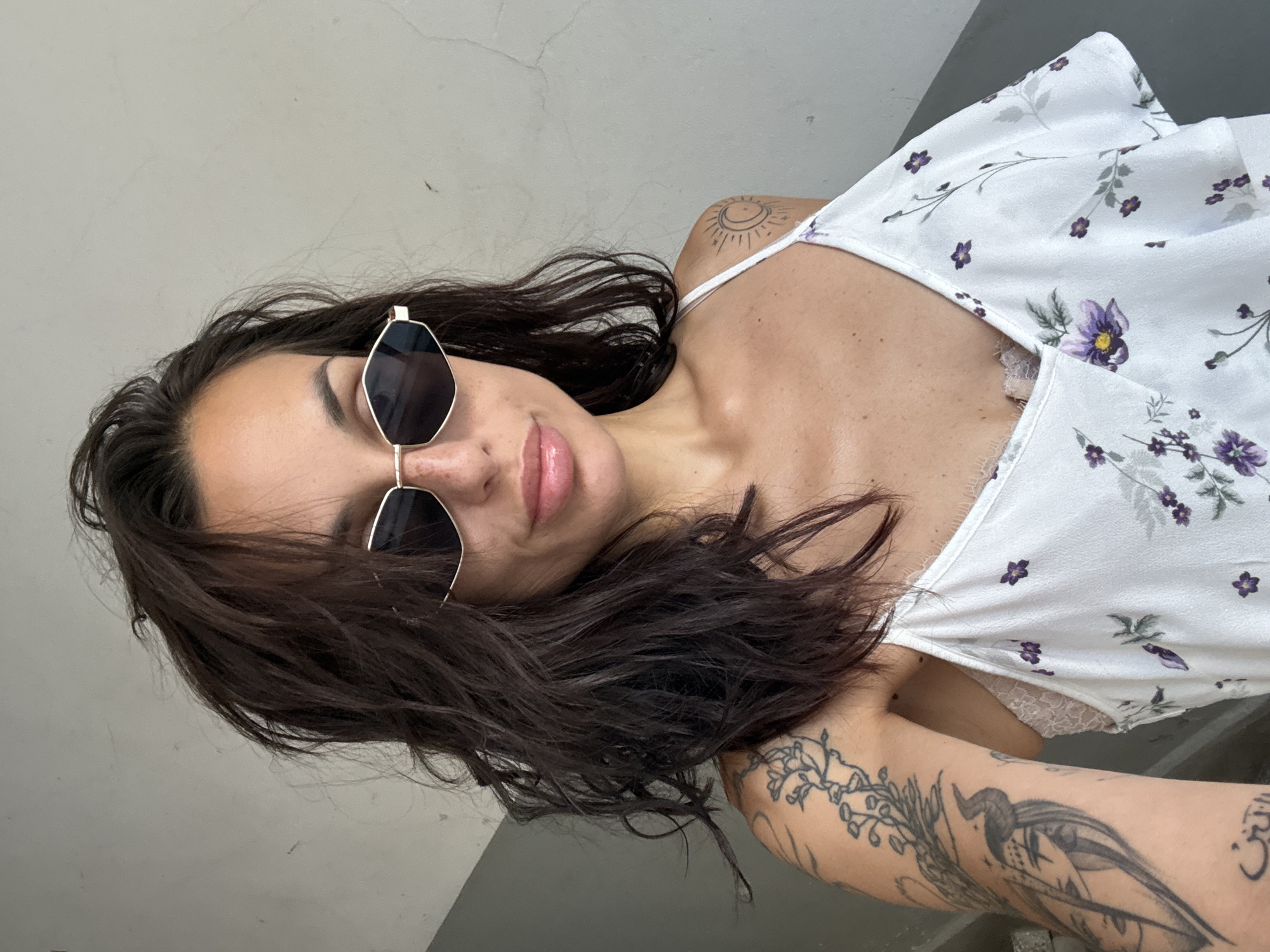 The image shows a medium shot of a woman posing for a selfie against a textured gray wall. She has dark, wavy hair that partially covers her face and shoulders, and she is wearing gold-rimmed, geometric sunglasses. Her lips are glossy and slightly parted, and there are a few freckles visible on her nose and cheeks. She is wearing a white camisole with a floral pattern of purple flowers and green leaves. A hint of lace from a bra is visible at the neckline. The woman has several tattoos: a large, detailed black and grey tattoo covering her left arm and shoulder, featuring botanical elements and what appears to be a bird or mythical creature. Another tattoo is visible on her right shoulder, depicting a crescent moon within a sun-like circle, done in a similar purple ink to the flowers on her dress. The background is a neutral, slightly weathered gray wall, suggesting an urban or indoor setting. The scene is set on a staircase in an apartment building in Pleven, Bulgaria.