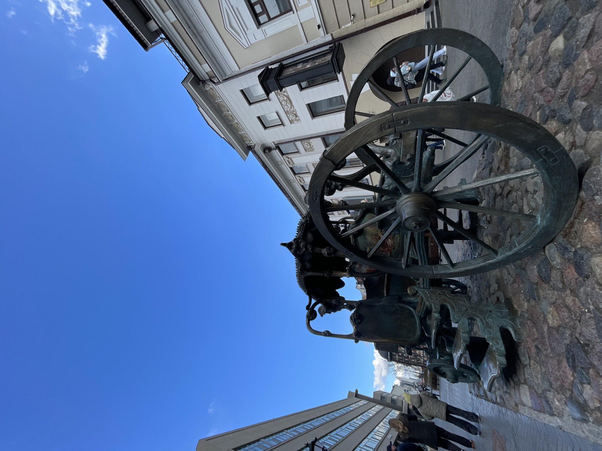 The image captures a vibrant daytime street scene, with a prominent bronze sculpture dominating the foreground. This large, ornate sculpture appears to be an elaborate carriage or cart, featuring sizable spoked wheels and intricate decorative elements. A person is visible sitting within the carriage section of the sculpture, suggesting its interactive nature as a monument or art installation.
The sculpture rests on a paved surface composed of both cobblestones and modern paving stones. In the background, buildings line the street. To the right of the sculpture stands an elegant classical-style building with white facades, arched windows, and decorative cornices. To the left, a more contemporary building with multiple horizontal window panes is visible. Several people are seen walking on the sidewalks and street, some partially obscured by the sculpture, indicating typical urban activity.
The sky is a clear and bright blue with a few scattered white clouds, signifying sunny weather. Given the location context of Kazan, Russia, the sculpture is identifiable as the famous "Kazan Carriage" or "Kazan Cat" monument. No discernible text is visible in the image.