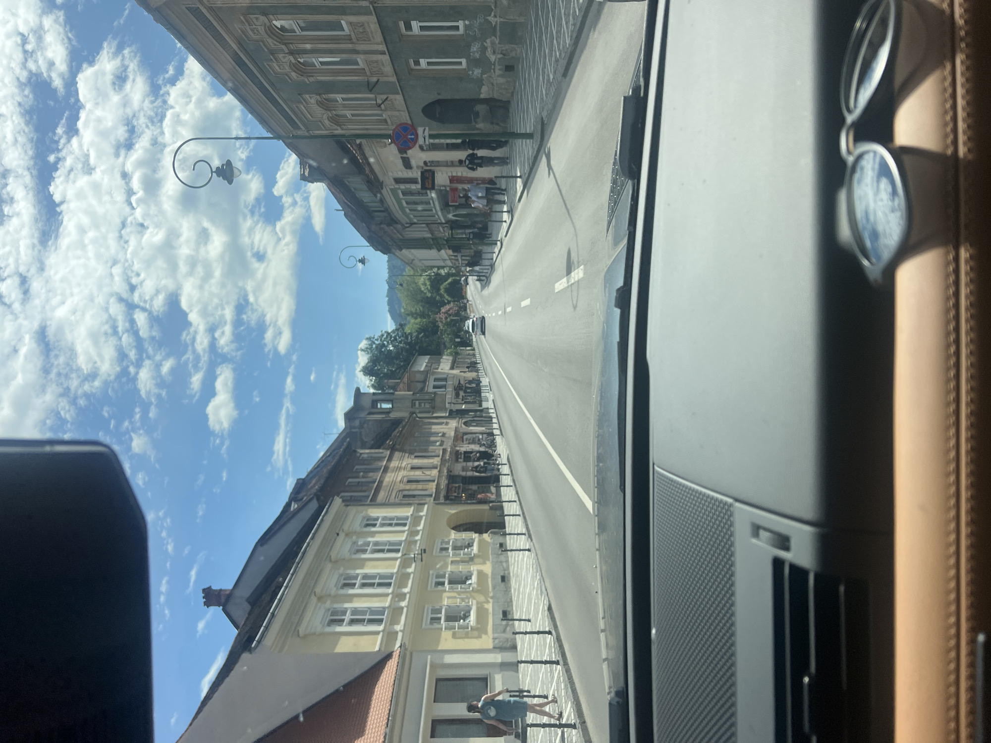 The media file depicts a street scene taken from inside a vehicle. The perspective is tilted, with the sky and buildings appearing to be on the sides of the frame.
The scene shows a street lined with buildings that have traditional architecture. The buildings are multi-story, with details like windows, balconies, and decorative elements. One building on the left has a weathered facade, while another on the right is painted yellow with white trim. The street itself is paved and has lane markings. There are streetlights with decorative scrolls. In the distance, there are trees and hills.
Several people are visible along the sidewalk. One person is walking on the sidewalk, appearing to be looking at their phone. Others are seen in small groups, possibly conversing or moving about. Some parked bicycles are also present.
The sky is blue with scattered, fluffy white clouds. The lighting suggests it is daytime, likely midday given the brightness. The weather appears to be pleasant.
Inside the vehicle, sunglasses are placed on the dashboard, and part of the car's interior, including a speaker grille, is visible.
No discernible text is clearly visible in the image.