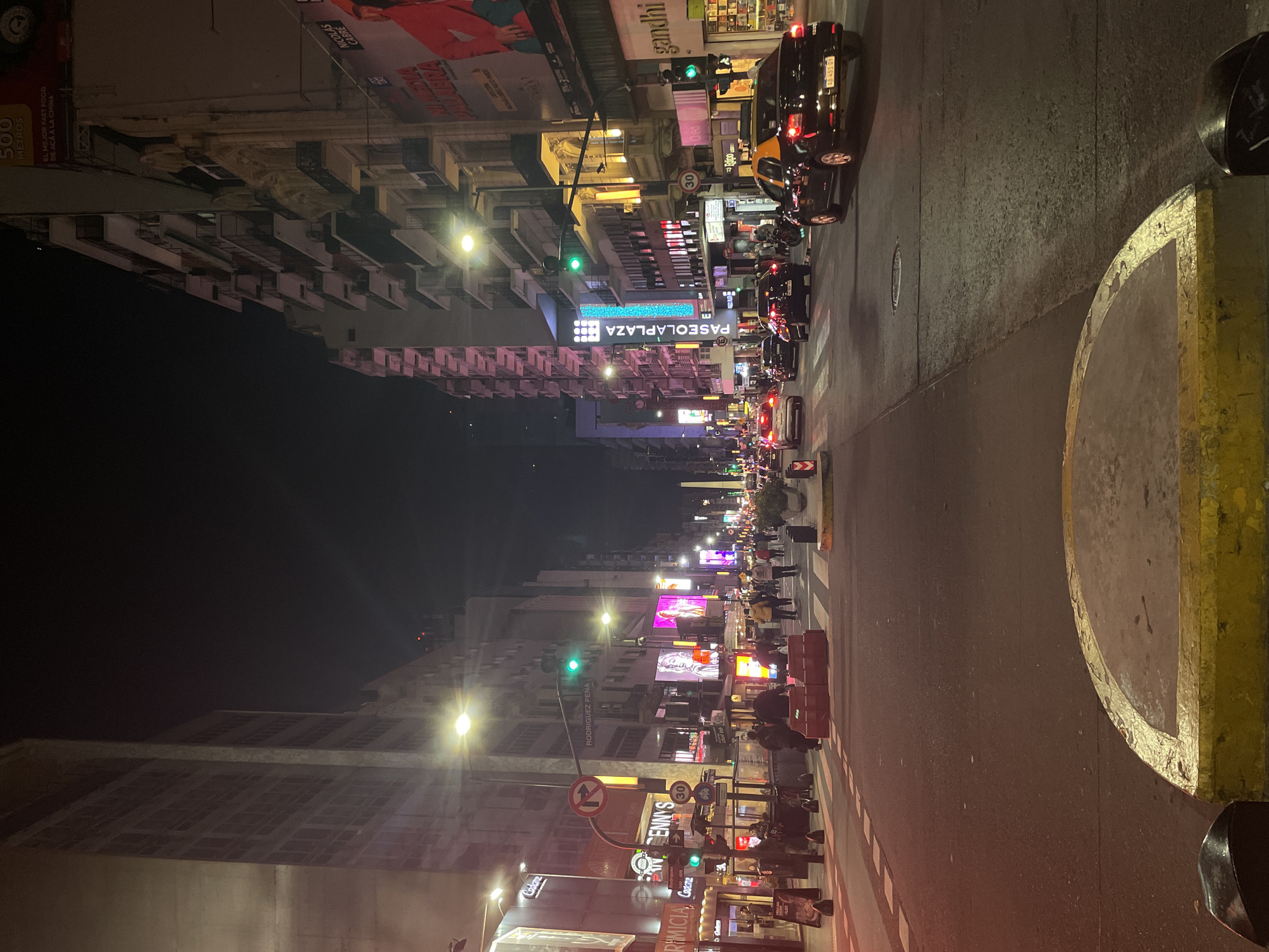 The image captures a night scene on Corrientes Avenue in Buenos Aires, Argentina. The avenue is illuminated by streetlights and neon signs, including a large "PASEOLAPLAZA" sign and various commercial advertisements. Traffic is visible on the road, consisting of cars, taxis, and motorcycles. Pedestrians are seen walking along the sidewalks, indicating a busy urban environment. The buildings lining the avenue are a mix of architectural styles, suggesting a historic and vibrant commercial district. Street furniture, such as trash bins and street signs, are visible along the sidewalk.